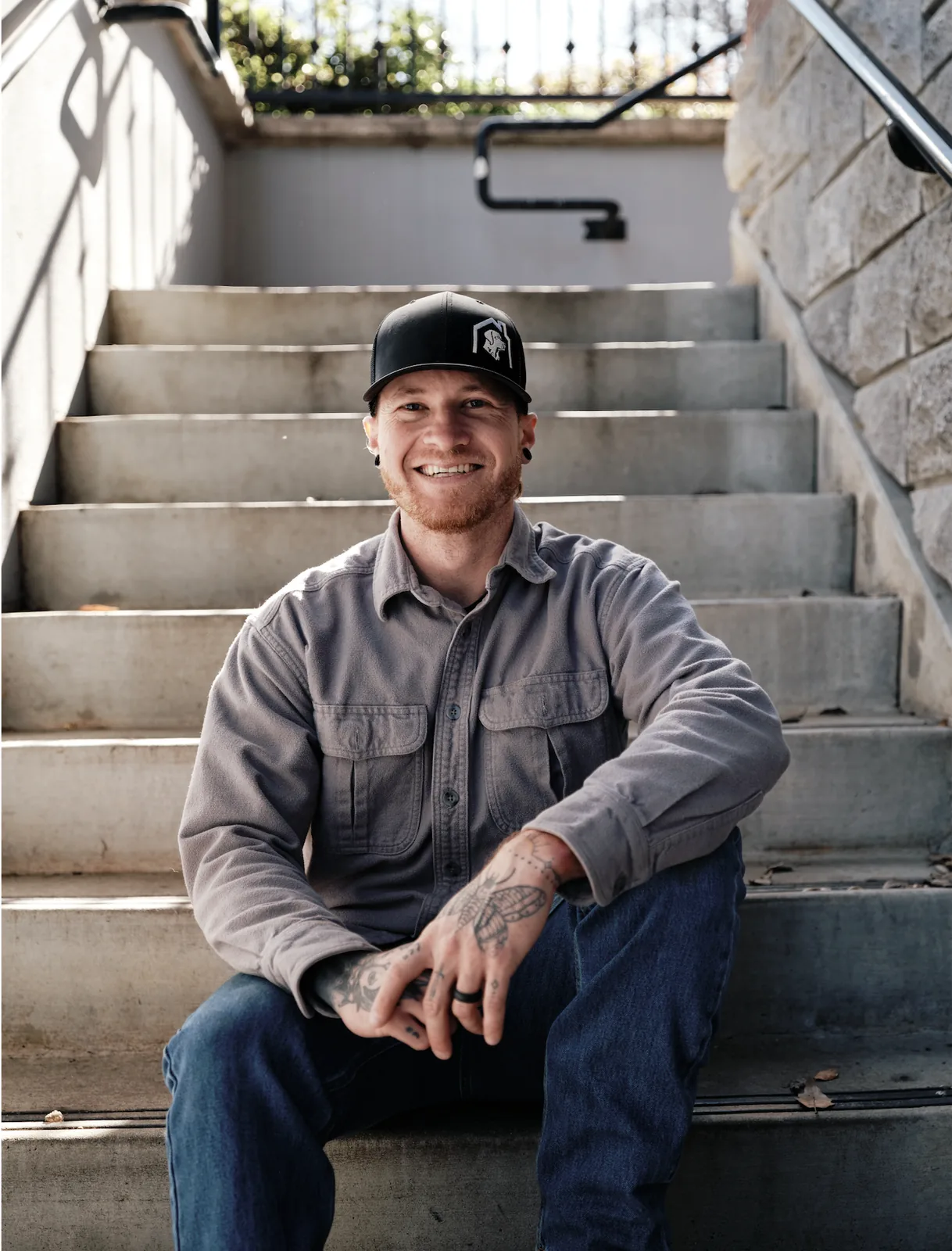 A smiling man with tattoos on his right arm, wearing a black baseball cap and a gray button-up shirt, sitting on outdoor concrete stairs with a stone wall on his right and a metal railing on his left, in daylight.