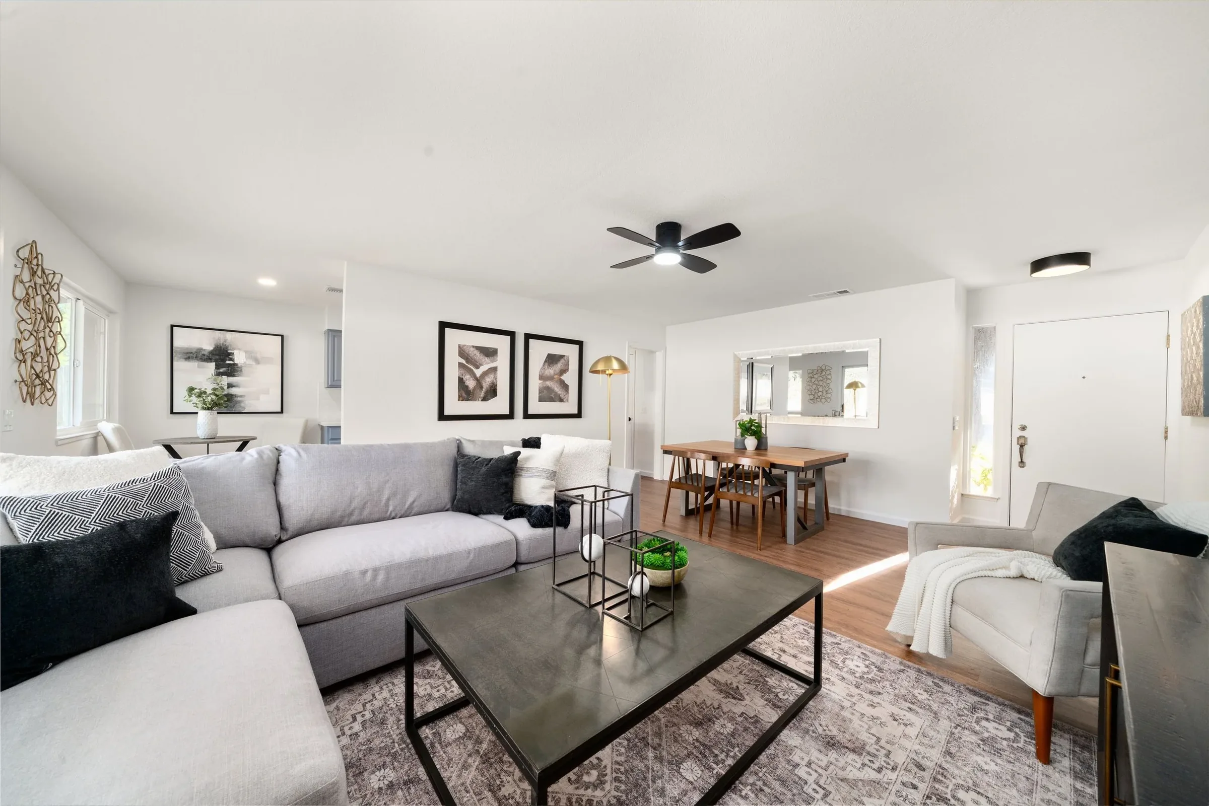 Living room with a gray sofa, black and white patterned pillows, black coffee table, white armchair, wall art, and a dining area with a wooden table and chairs, decorated with potted plants, in a bright, modern home.
