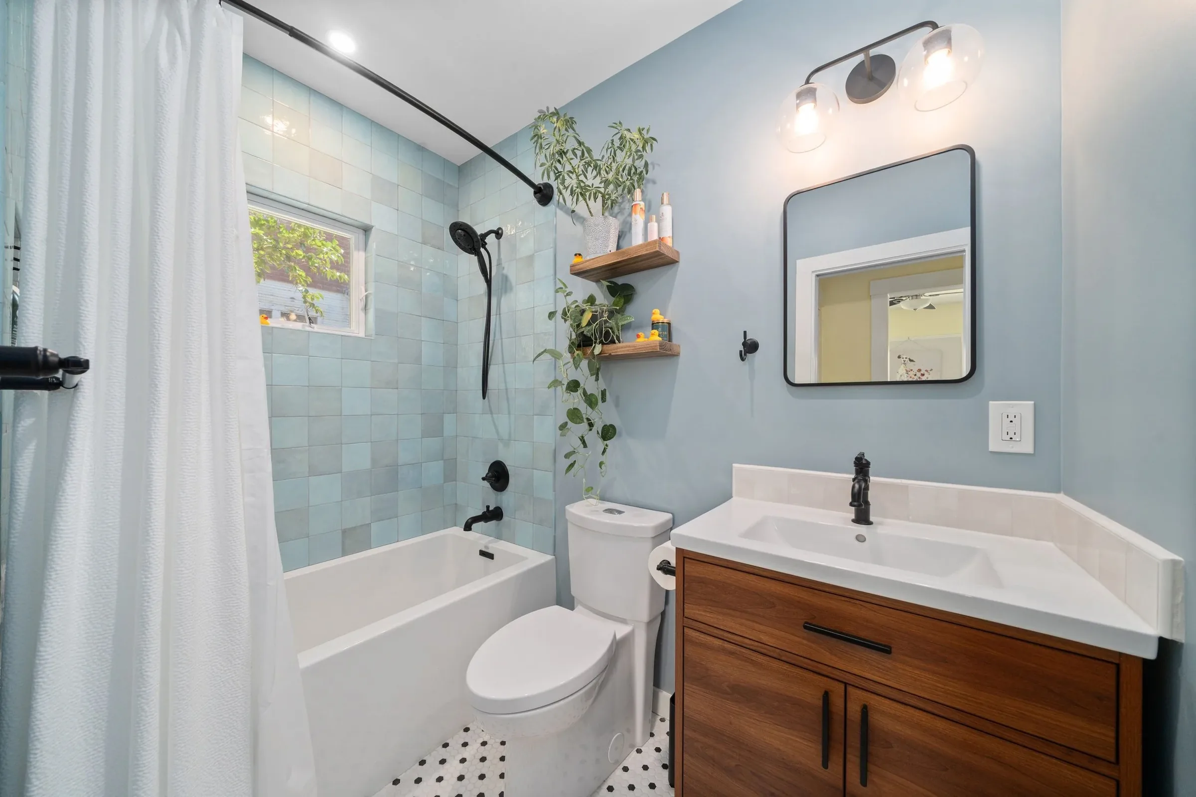 Modern bathroom with a bathtub/shower, a white toilet, a wooden vanity with a white sink, a mirror, and wall-mounted lights. There are two wooden shelves with plants and toiletries, a window with curtains, and a black showerhead with a rod.