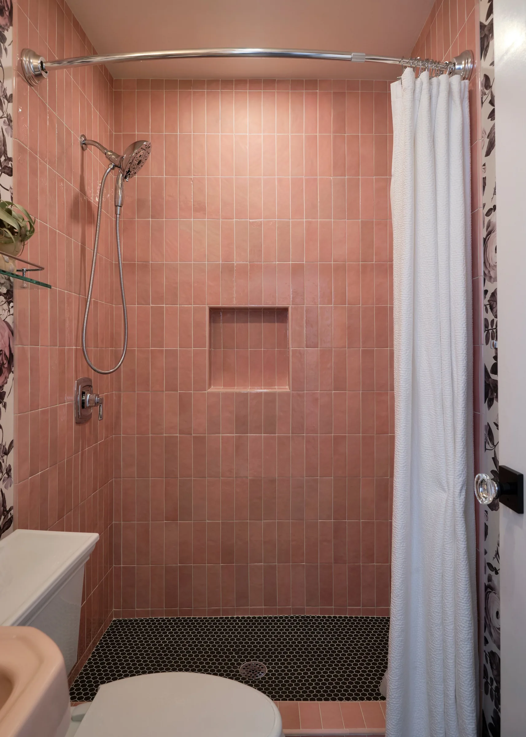 A small bathroom with pink tiled walls, a shower area with a curved metal shower rod and a white curtain, a built-in shower niche, black hexagon floor tiles, and a toilet in the foreground.