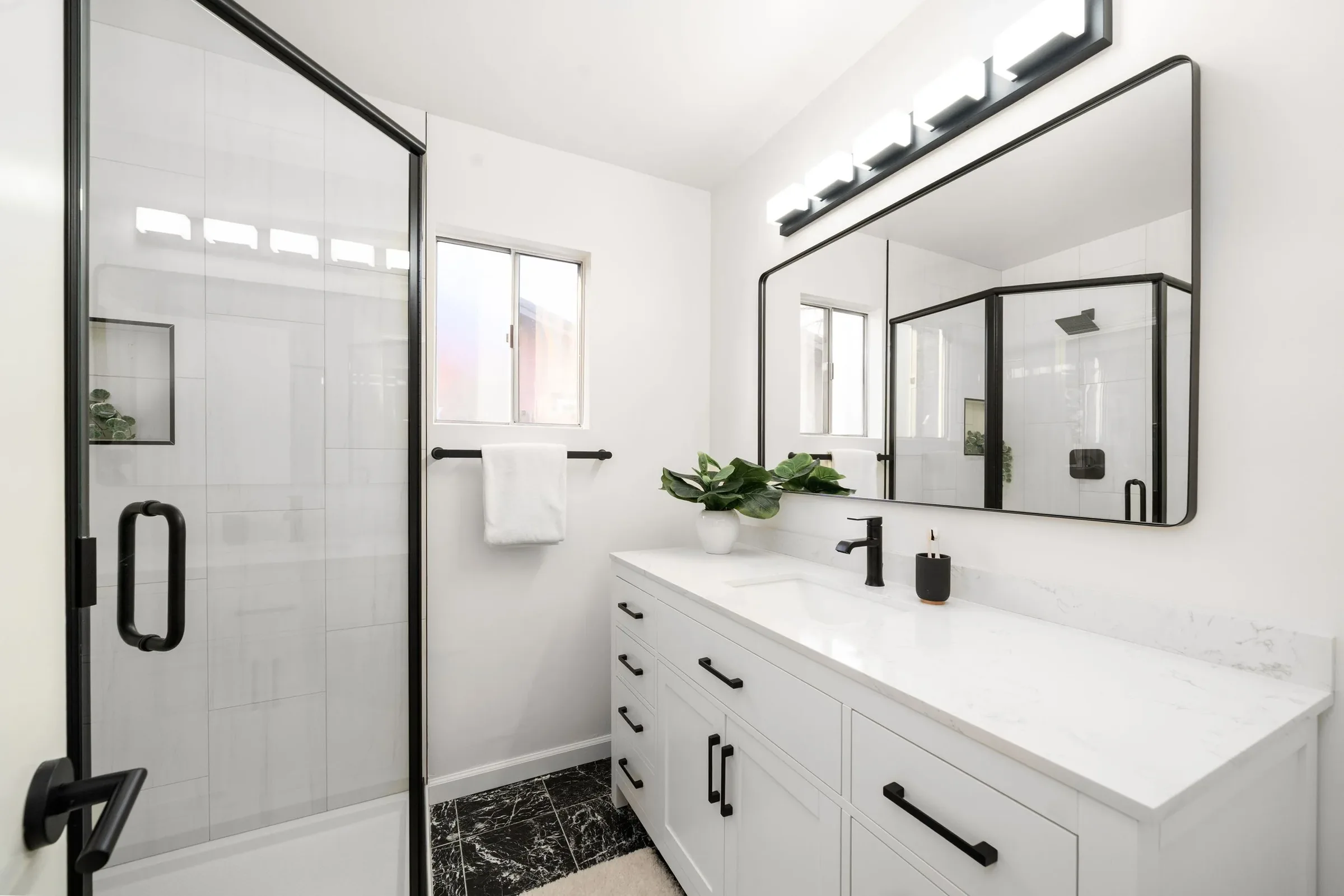 Modern bathroom with white vanity, black fixtures, large mirror, glass shower enclosure, and black and white marble floor.