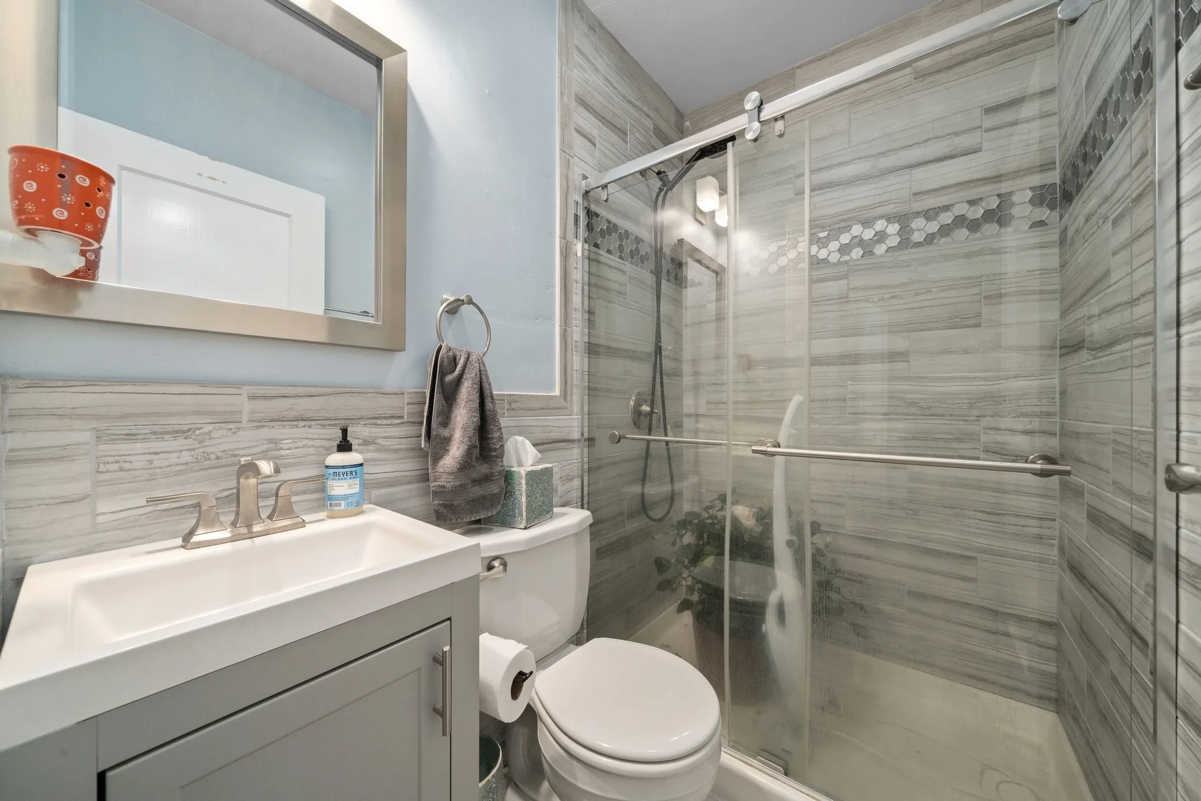 A small bathroom with a light blue painted wall and marble-look tiles. There is a white vanity with a sink, a mirror above it, and a soap dispenser. A toilet with a toilet paper roll is next to the vanity. There is a glass-enclosed shower with a slid