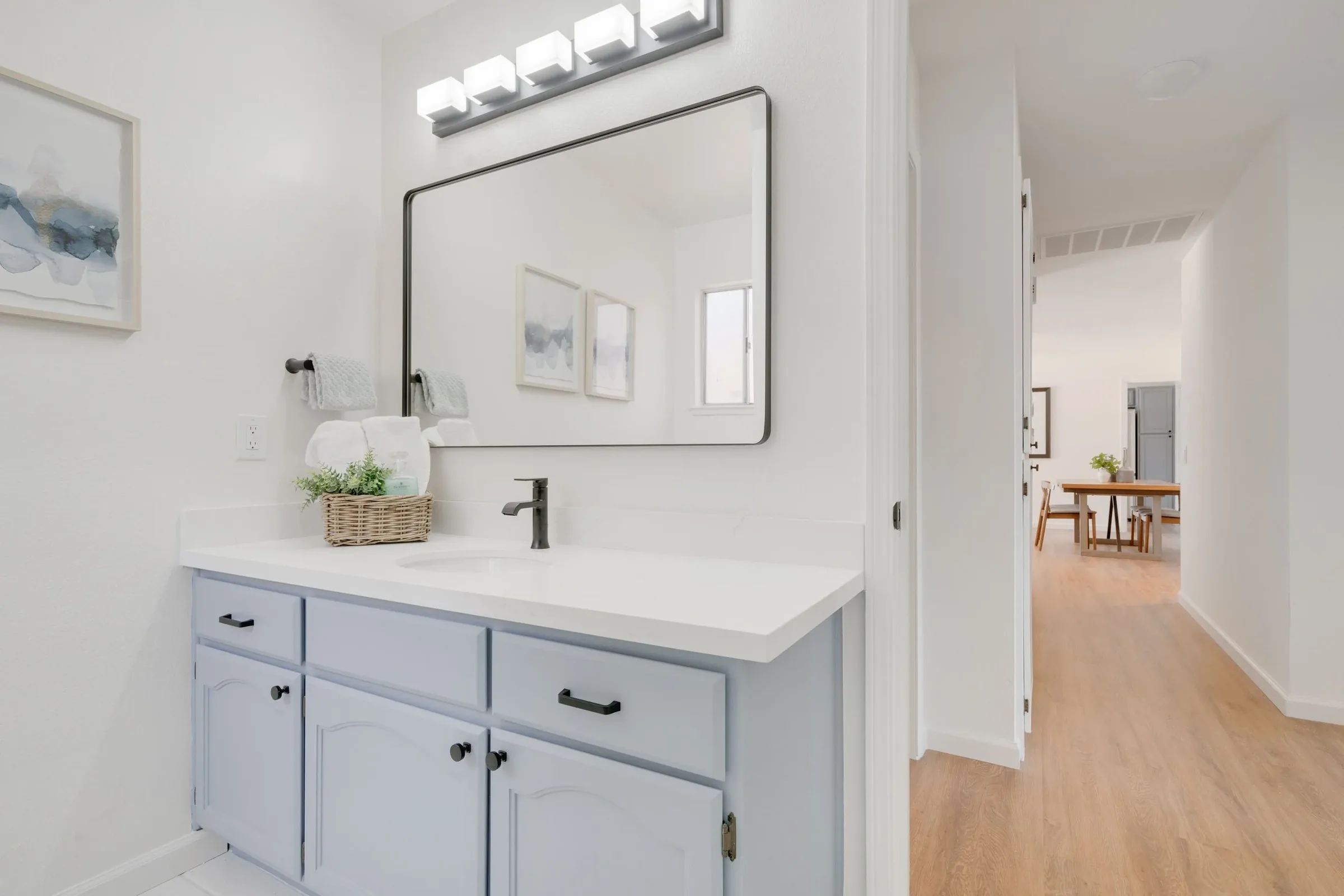 A bathroom vanity with a white countertop, a black faucet, and light blue cabinets. Above the vanity is a large rectangular mirror with a black frame, and above the mirror is a set of five square white light fixtures. To the left of the mirror are two abstract watercolor art pieces in white frames. There are two white towels hanging on a black towel bar, and a small basket with toiletries and a green plant on the countertop. In the background, part of an open living area with a wooden table and chairs, light-colored walls, and a window is visible.