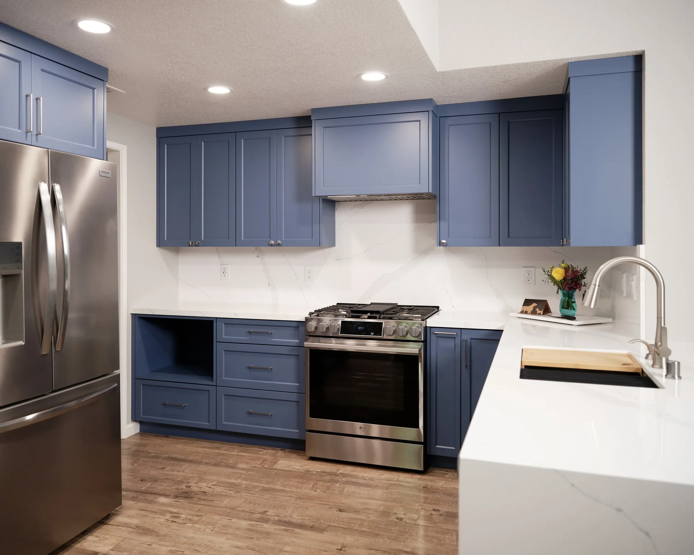 Kitchen with blue cabinets, stainless steel refrigerator and oven, white marble countertops and backsplash, and a small flower arrangement on the counter.
