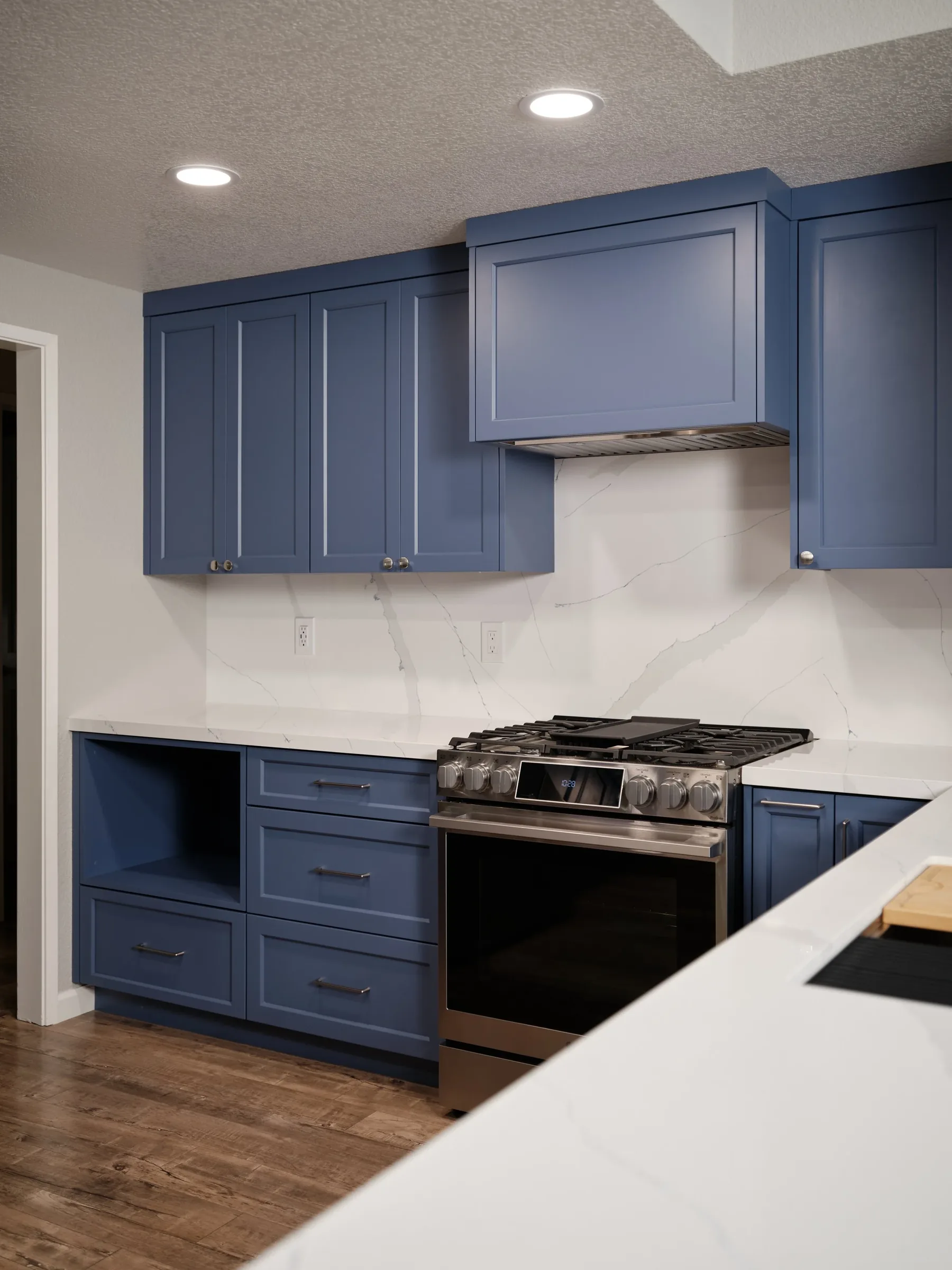 Kitchen with blue cabinets, white marble backsplash, stainless steel stove, and wooden flooring.
