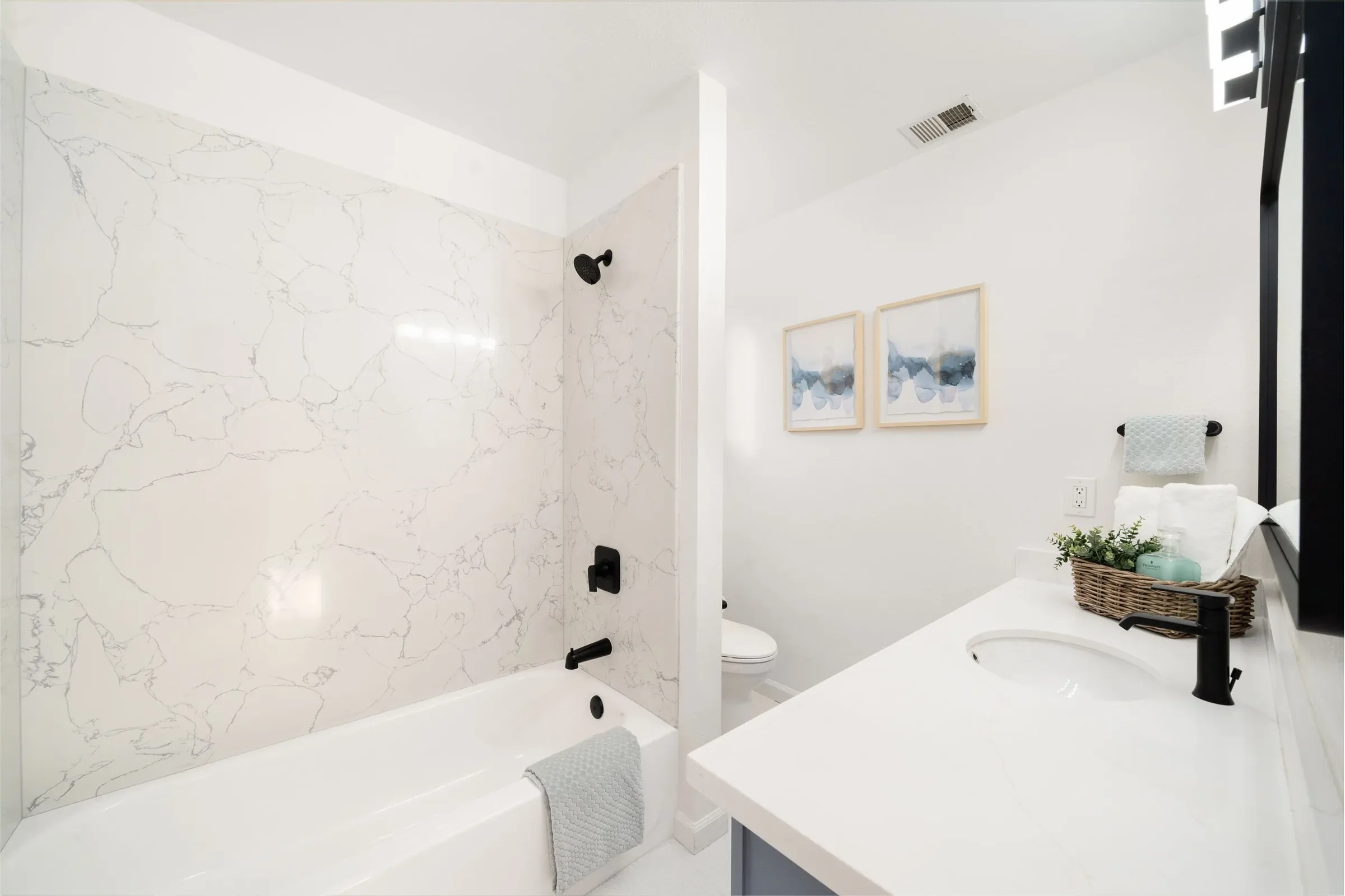 Modern bathroom with white marble shower wall, black shower fixtures, toilet, and vanity area with a basket of toiletries, framed artwork, and a towel.