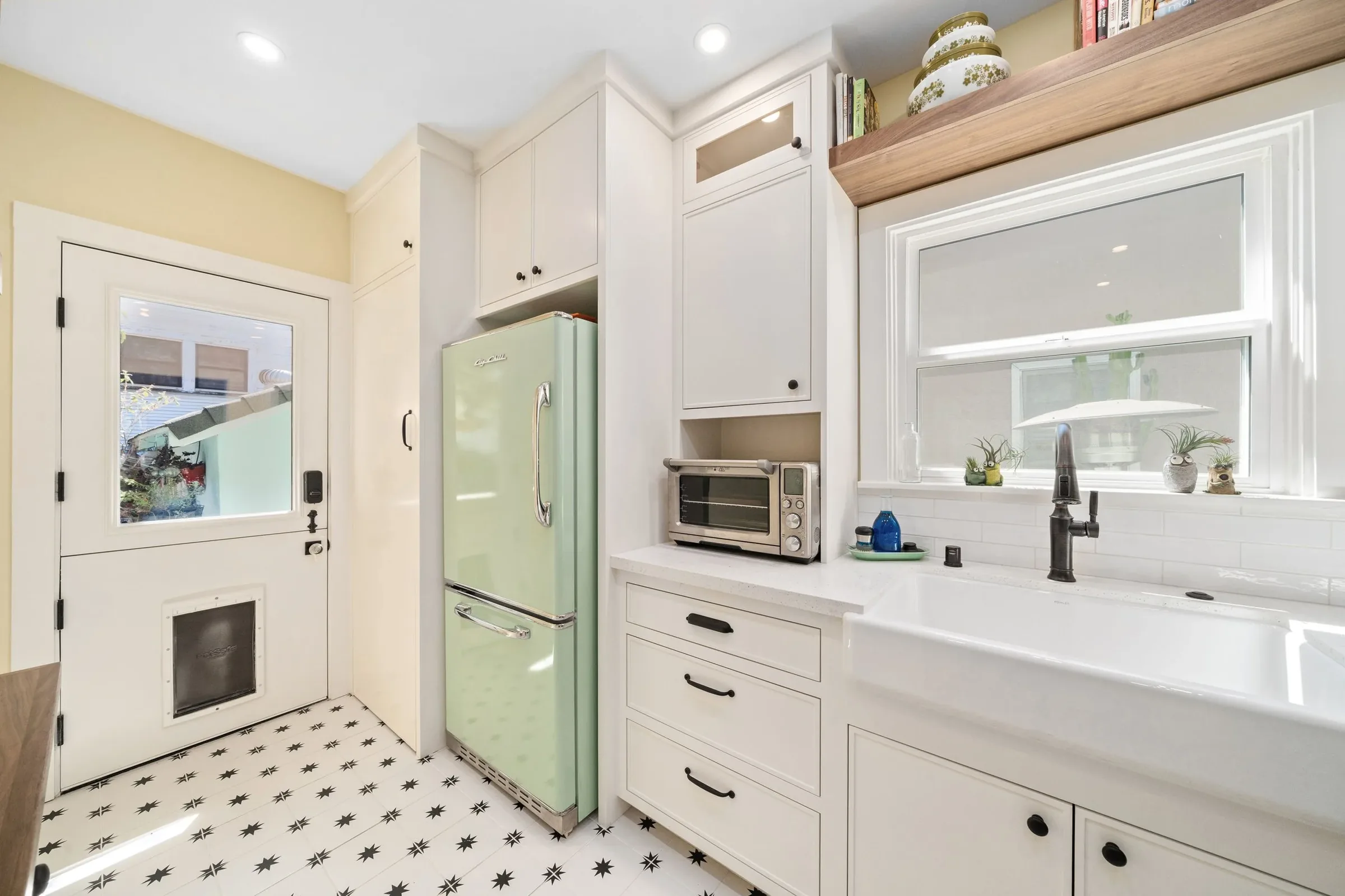 A bright kitchen with white cabinets, a black and white star-patterned floor, a light green vintage refrigerator, a microwave, a window with a view of a patio, and small decorative plants on the windowsill.