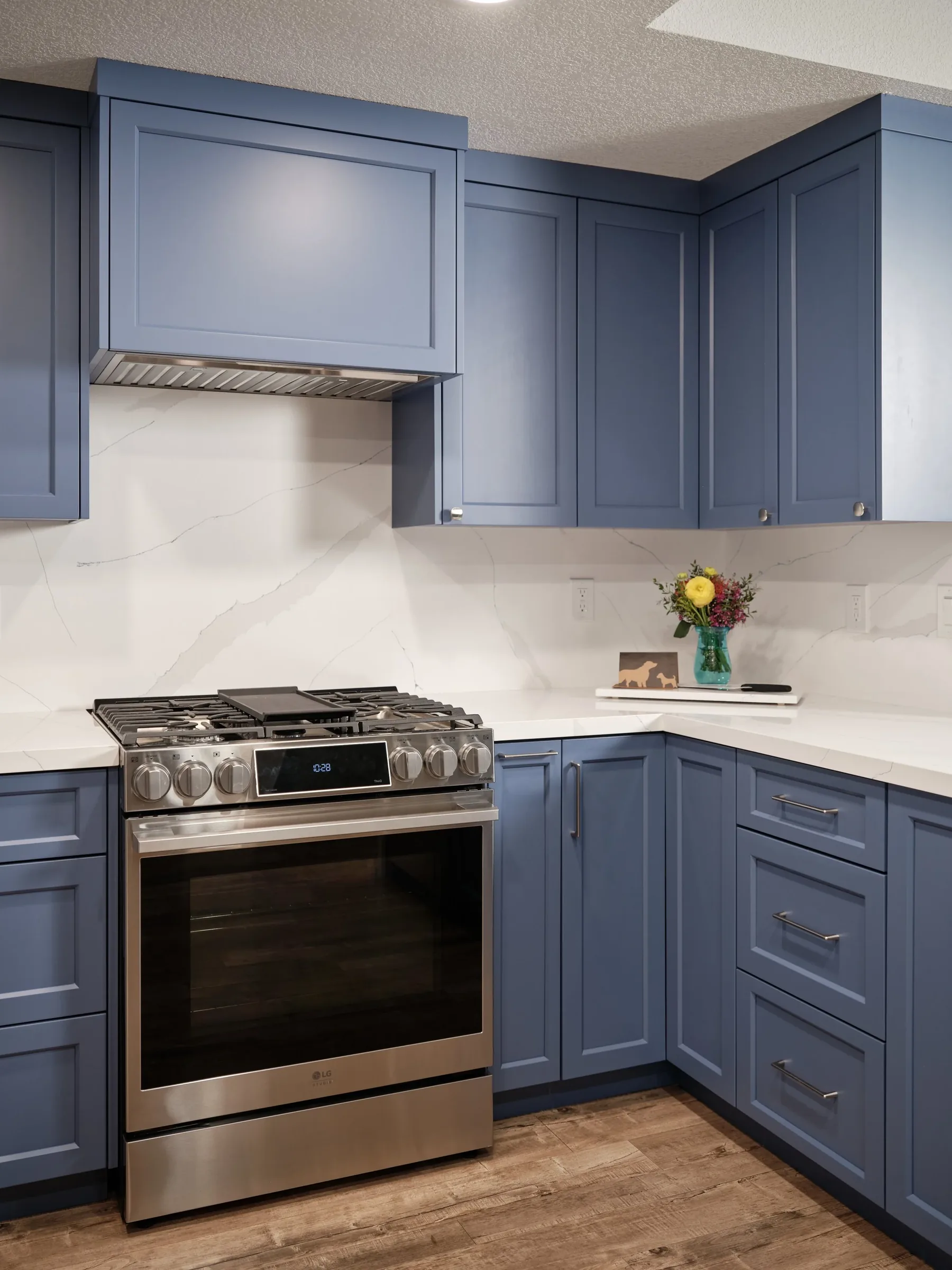Kitchen with blue cabinetry, stainless steel stove, white marble countertops with gray veining, and a bouquet of flowers in a teal vase.