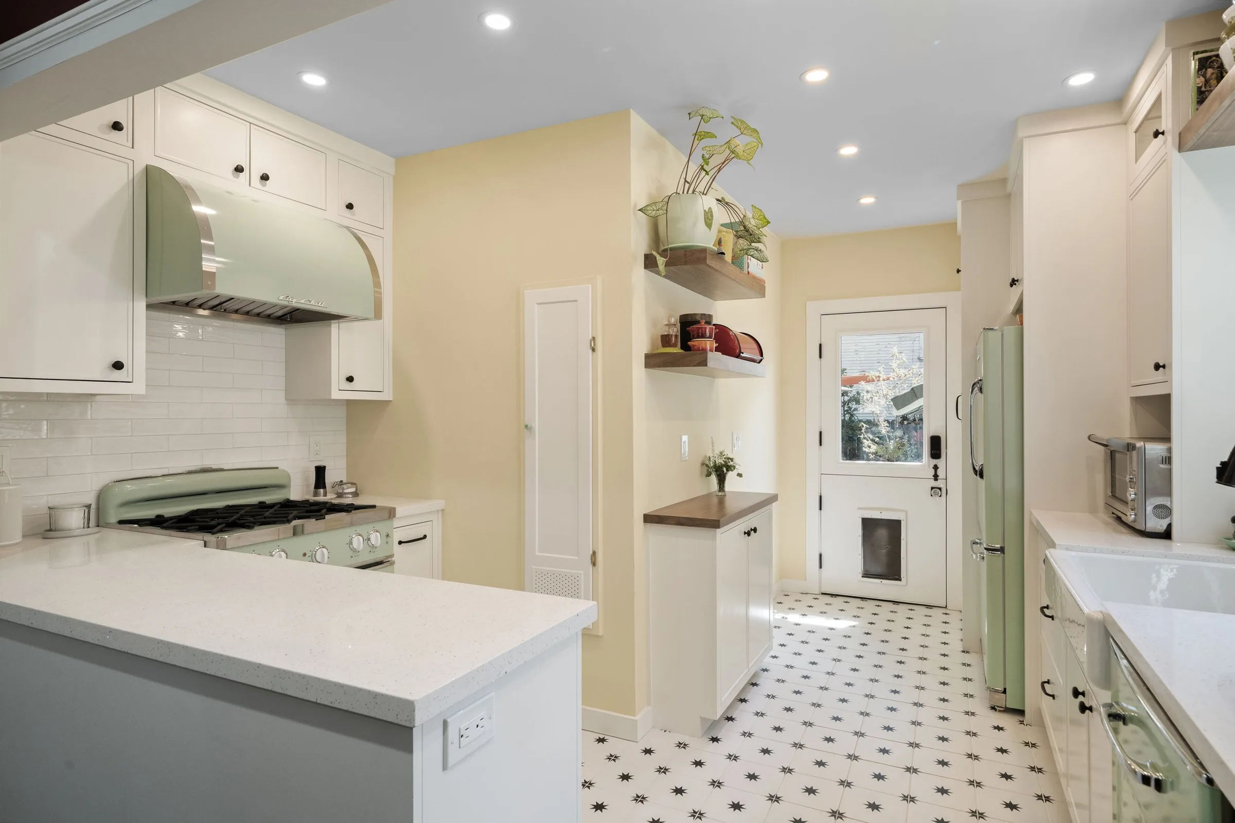 A bright kitchen with white cabinets, a stove with a light green range hood, and a window with a view outside. There are two floating shelves with plants and dishes, and a door at the back leading outside. The floor is decorated with black star patterns on a white background.