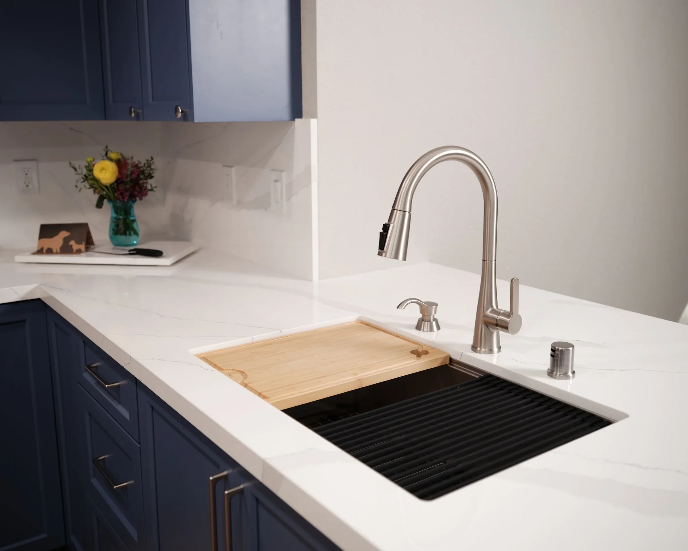 Modern kitchen with navy blue cabinets, white marble countertop, a stainless steel faucet, a wooden cutting board, a vase with yellow and pink flowers, and a black stovetop.