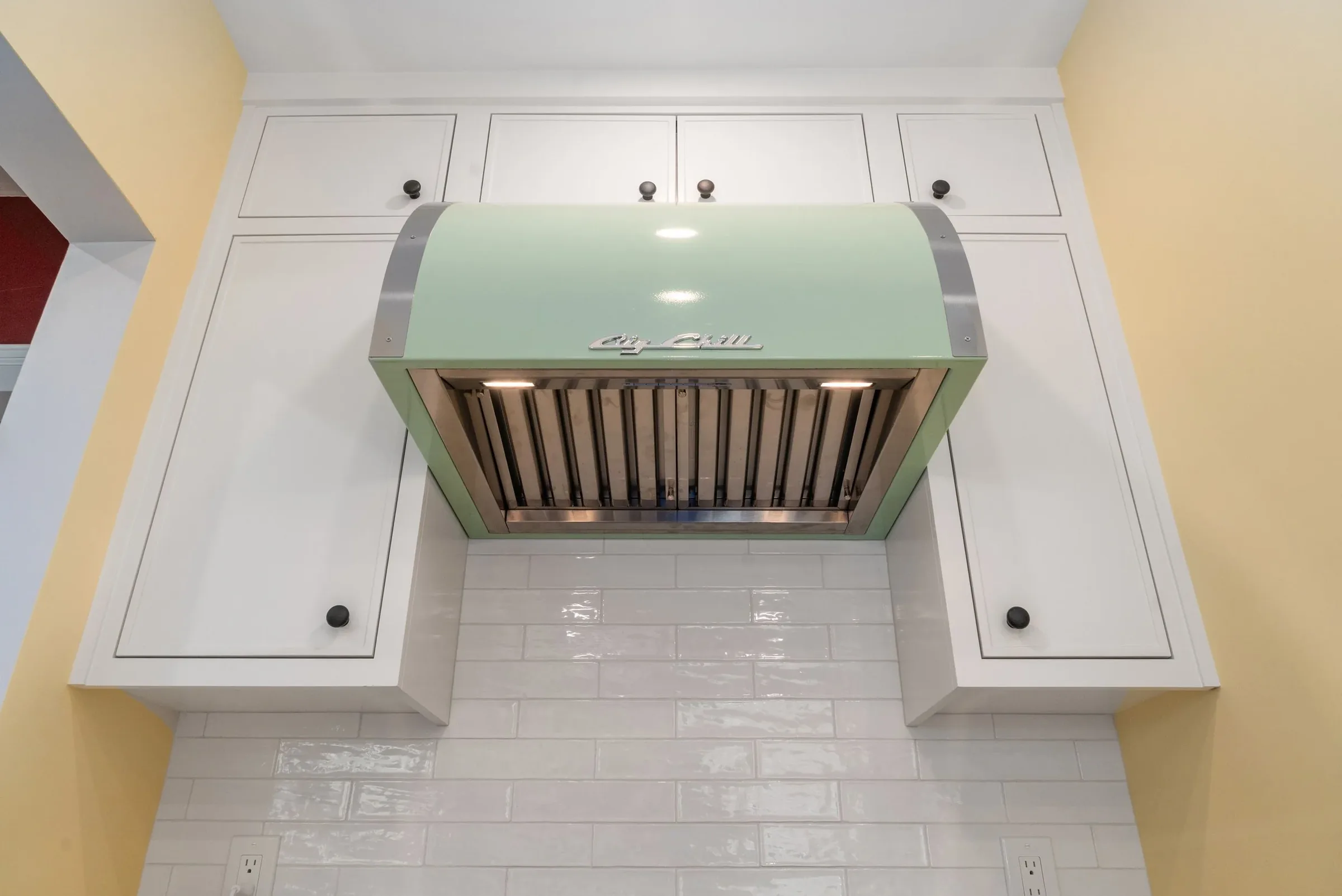 A kitchen range hood with a pale green front and stainless steel trim, mounted above a white tiled wall. White cabinets with black knobs are visible above the range hood, and yellow walls are on either side. A white electrical outlet is at the bottom