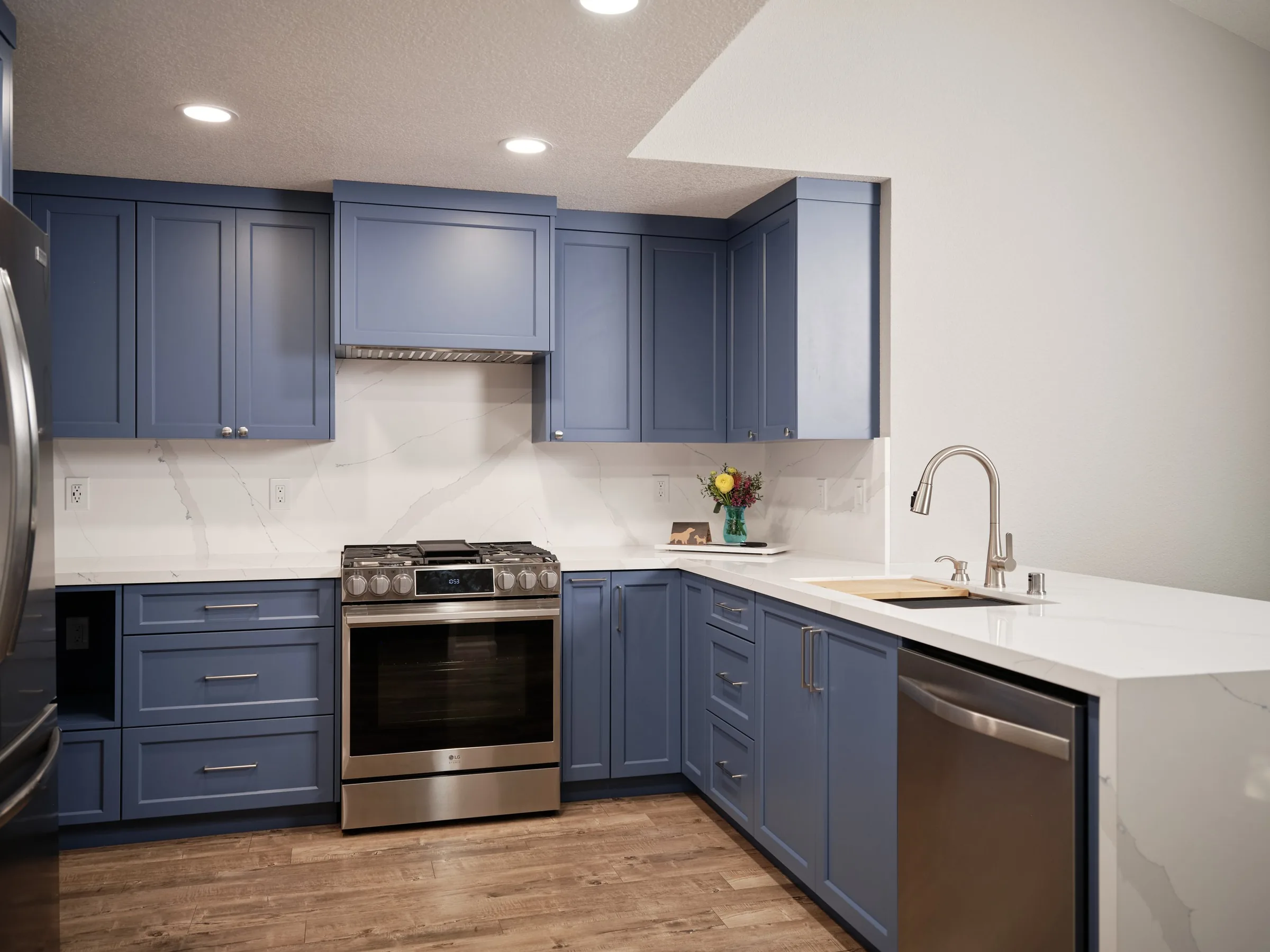 A modern kitchen featuring blue cabinets, a stainless steel oven and stove, a white marble countertop with subtle gray veining, and a small bouquet of flowers in a teal vase on the counter. The kitchen has a stainless steel dishwasher, and there is a