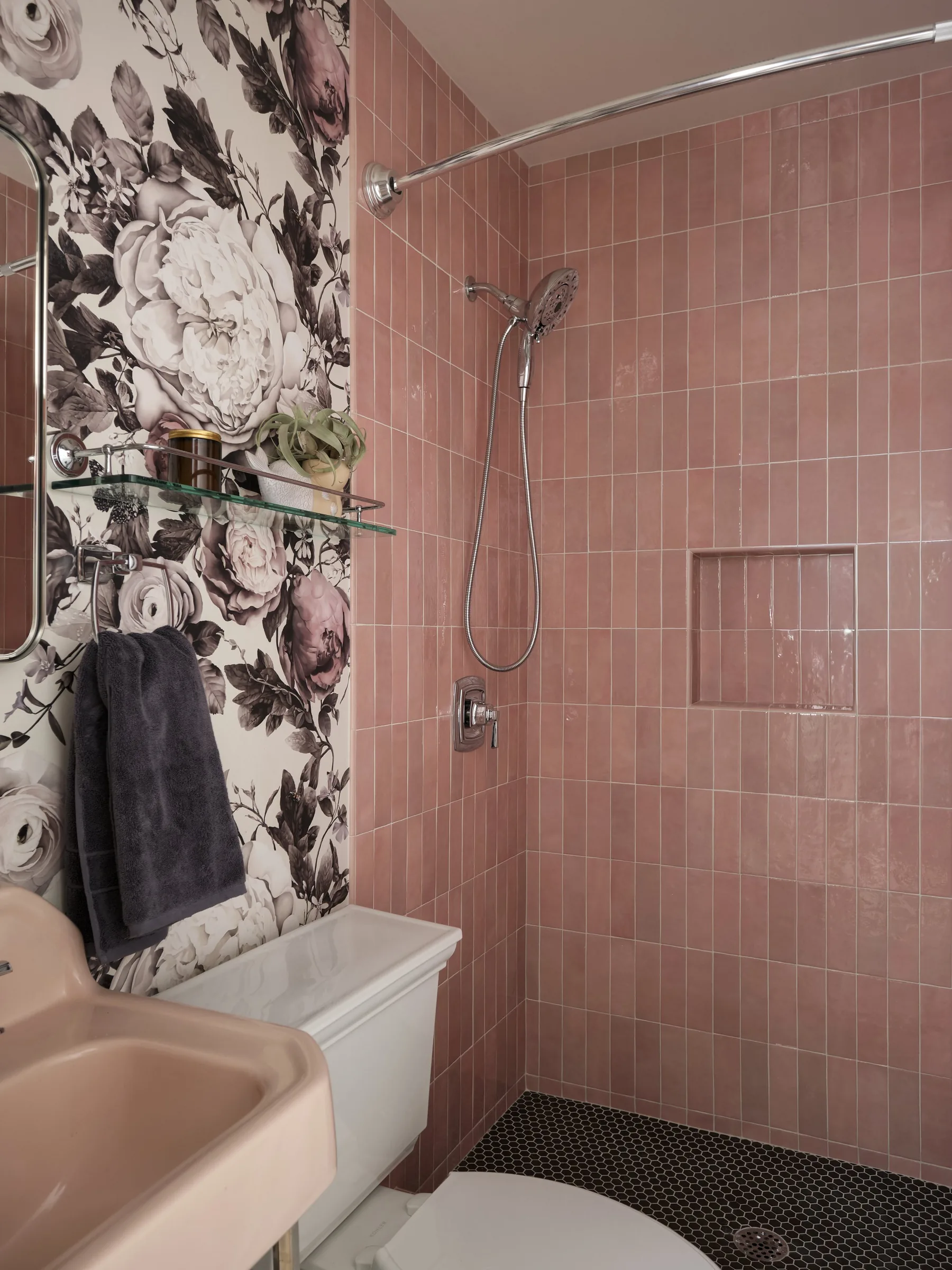 A small pink shower with pink tiled walls, a curved shower rod, a hand-held shower head, a wall niche, and a decorative floral wallpaper on one side. There is a black towel hanging on a bar and a small white shelf with plants and cosmetics.