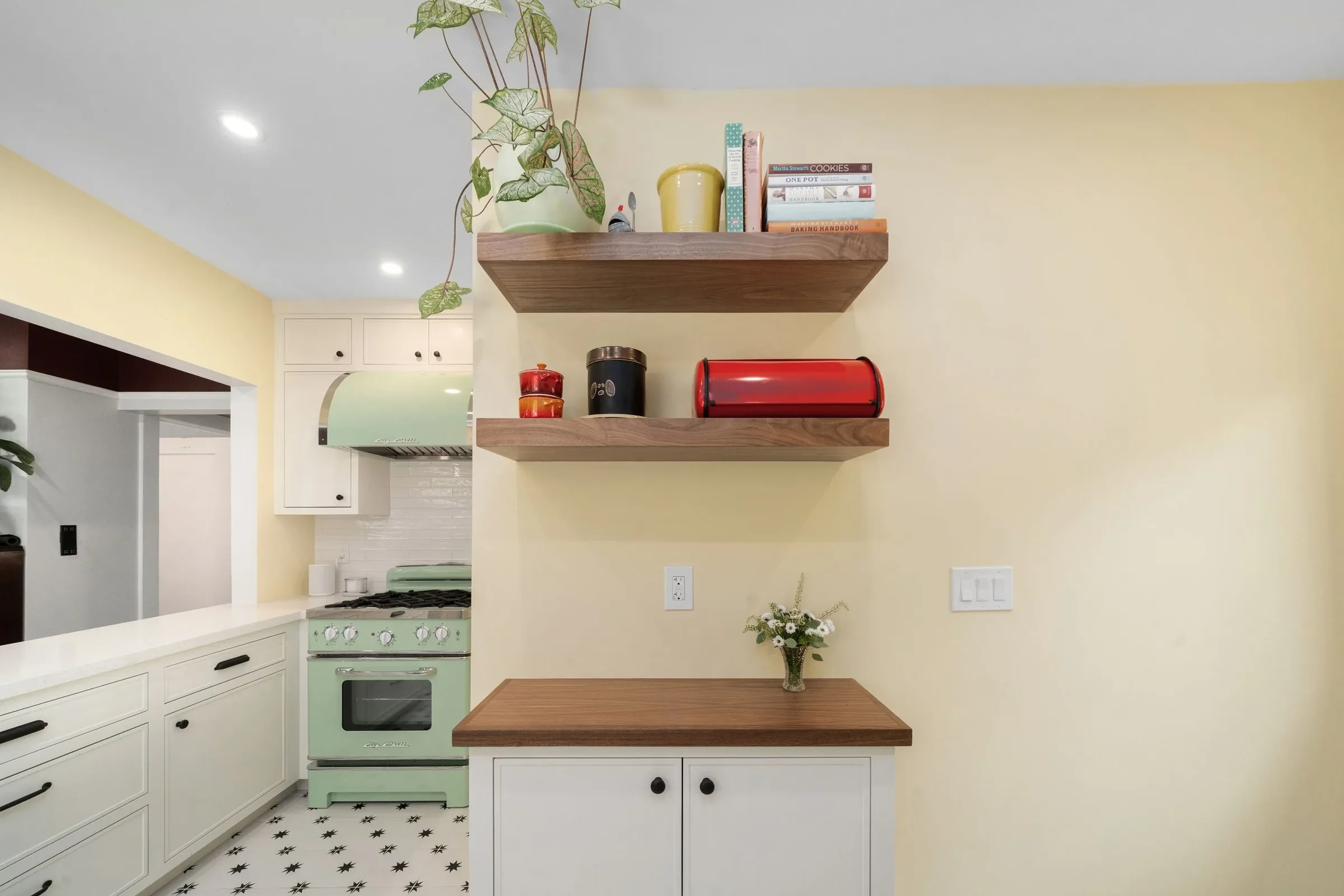 Kitchen with white cabinets, a mint green vintage oven, and two wooden floating shelves decorated with a plant in a cream pot, books, and kitchen accessories. There is a vase with white flowers on the countertop.