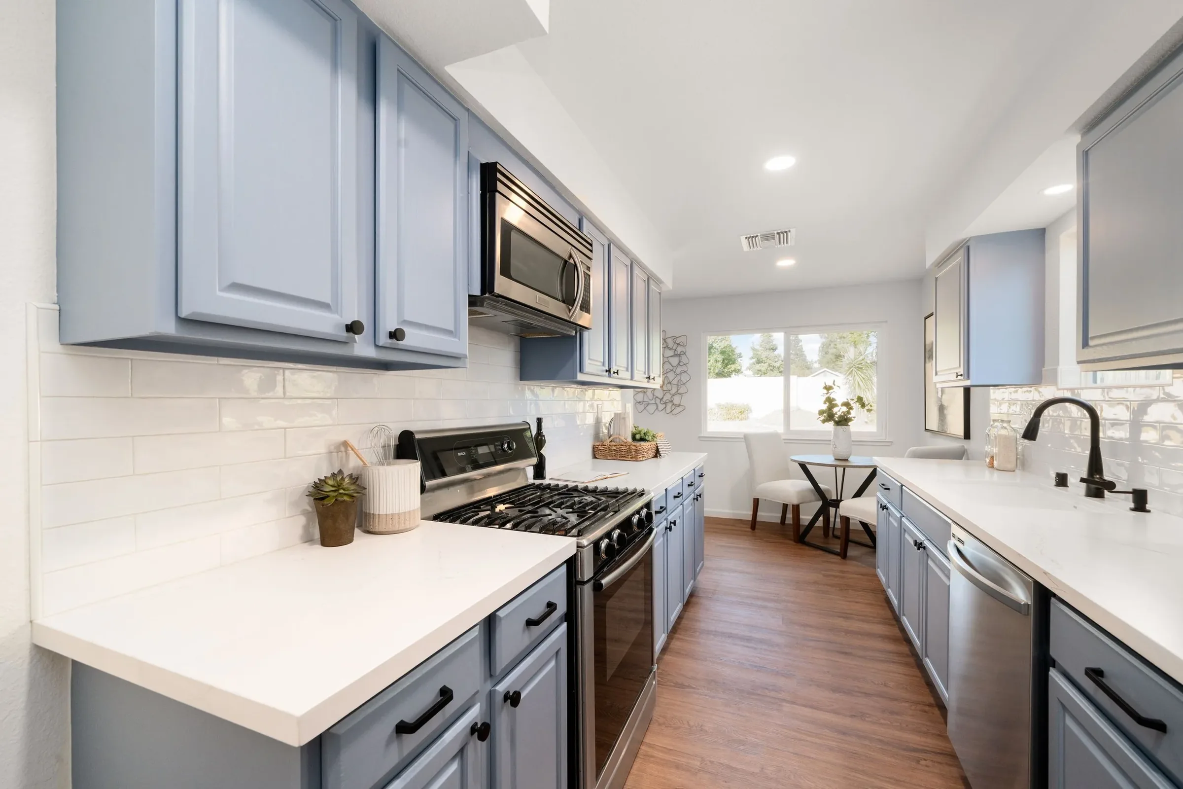 Bright kitchen with light blue cabinets, stainless steel microwave, white countertops, and a white tile backsplash. A window lets in natural light, showing outdoor greenery. Small decorative plants and kitchen utensils are on the counters, with a sta