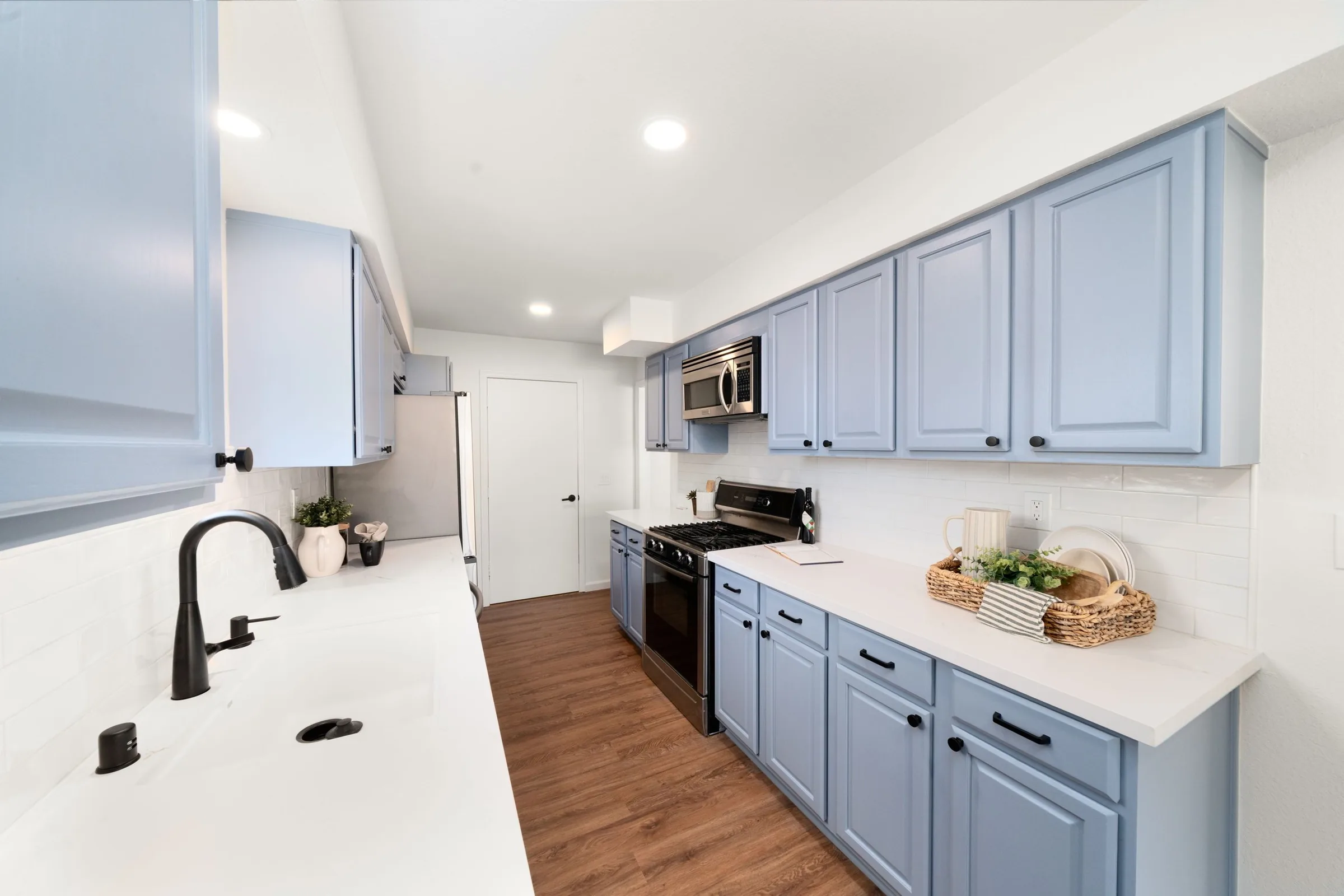 Kitchen with blue cabinets, white countertops, stainless steel microwave, oven, black faucet, and wooden floors.