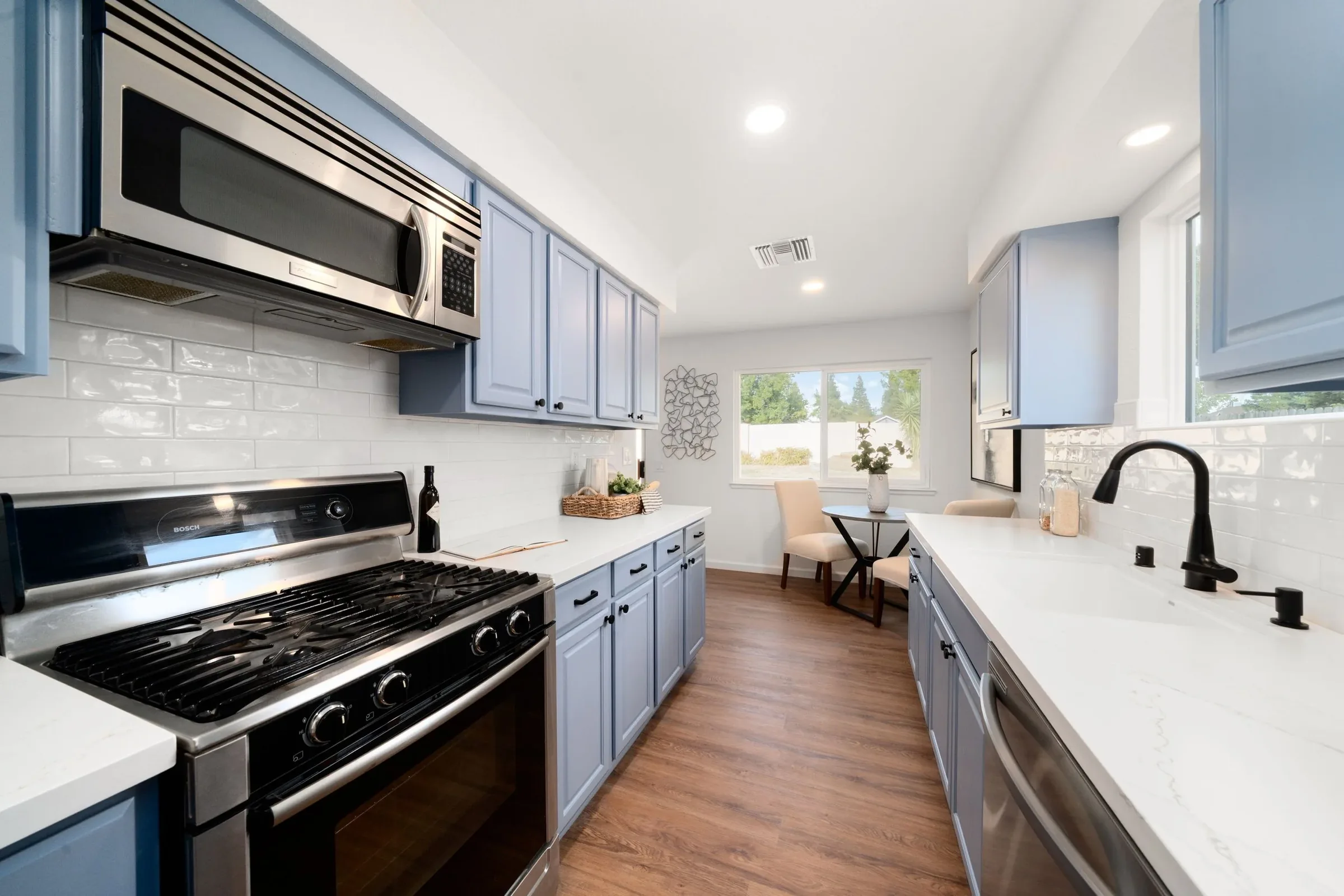 Kitchen with blue cabinets, stainless steel microwave, stove, and sink, with a window and dining area in the background.