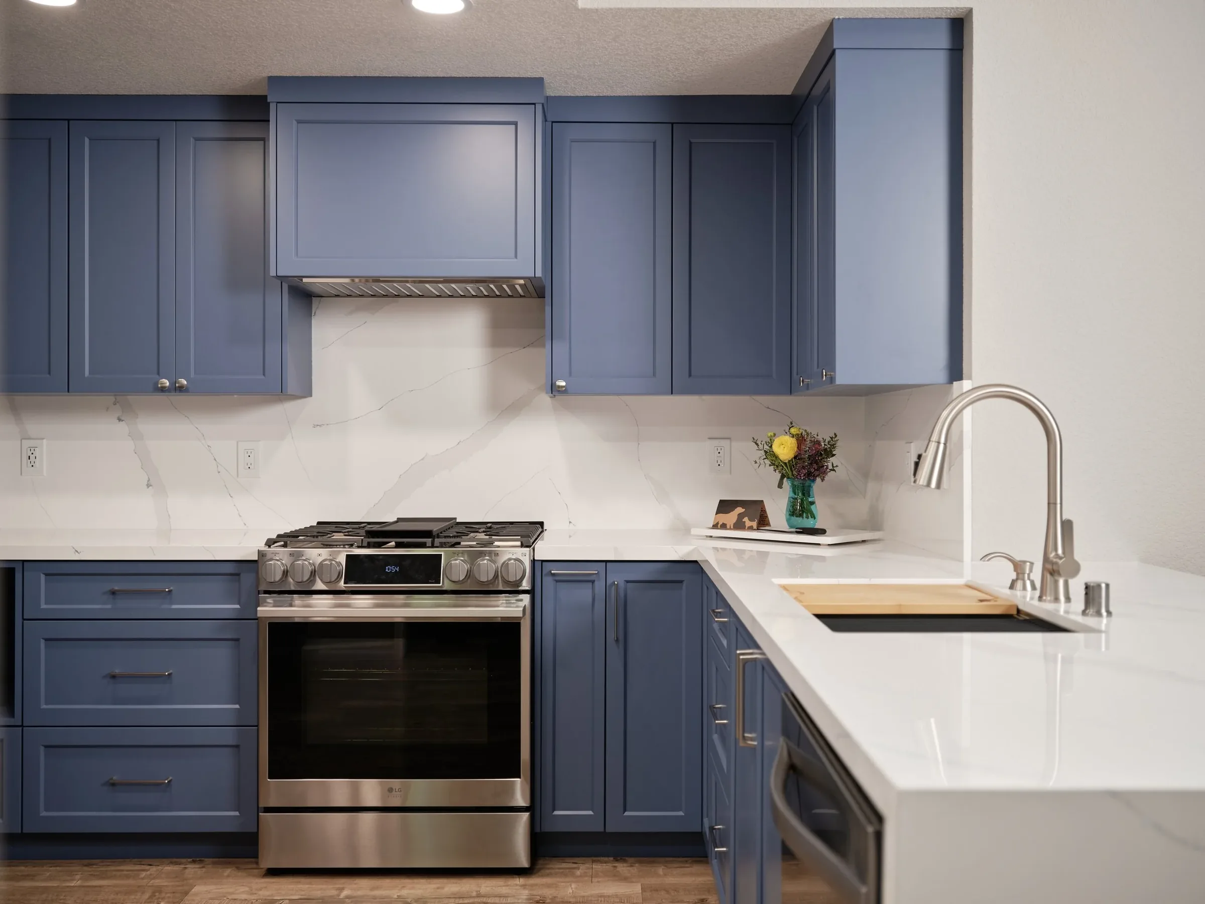 A modern kitchen with blue cabinets, a stainless steel oven and a white marble countertop. There is a vase with flowers on the counter and a stainless steel faucet over the sink.