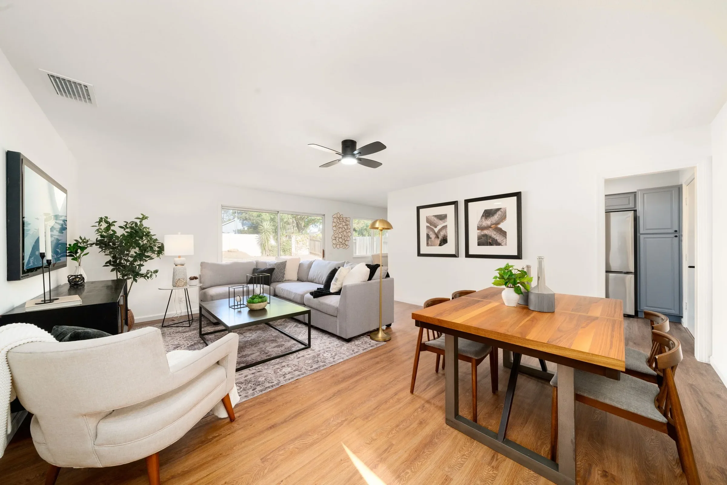 Living room with white walls, hardwood flooring, a gray sectional sofa, a white armchair, a black coffee table, artwork on the walls, a gold floor lamp, a flat-screen TV, and a window with greenery outside.