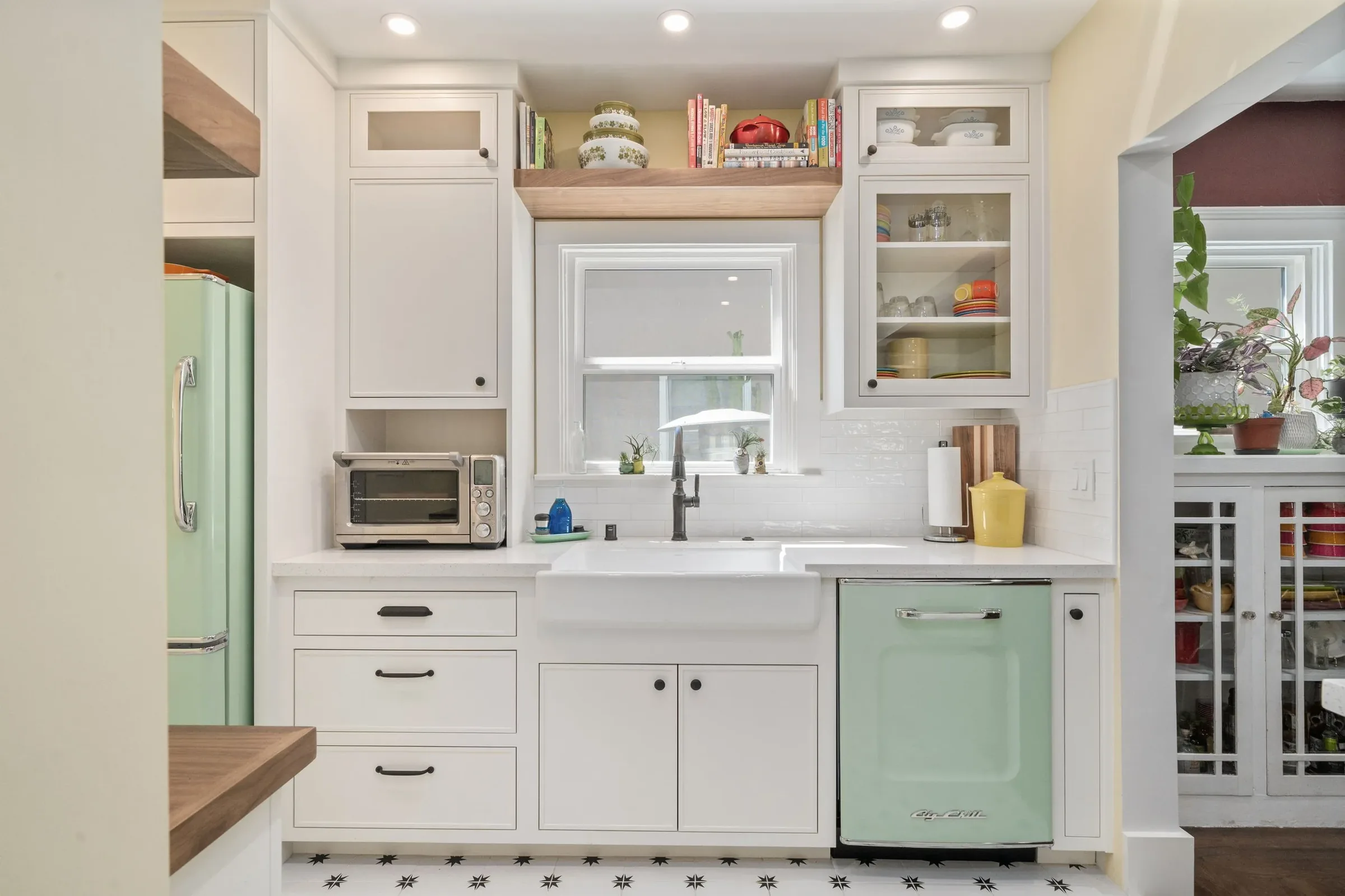 Kitchen with white cabinets, a white window above the sink, and a black star-patterned tile floor. Contains a pastel green refrigerator, a toaster oven, and decorative plants.