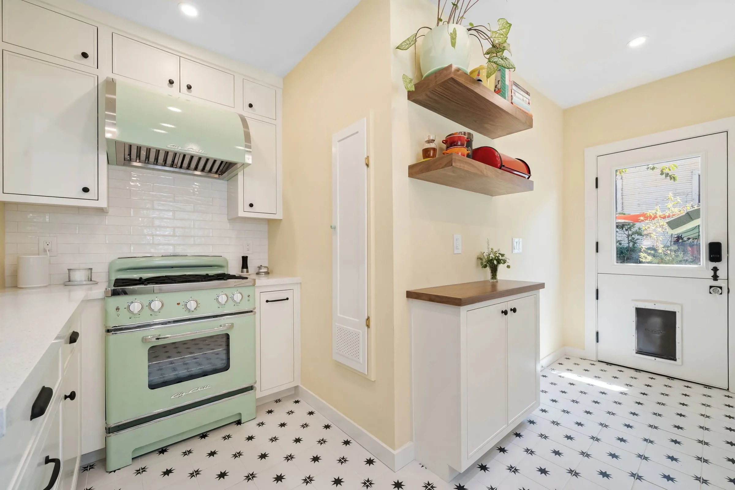 A kitchen with vintage mint green stove, white cabinets with black knobs, white subway tile backsplash, two floating wooden shelves with plants and cookware, a windowed door with a view outside, and black and white star-patterned floor tiles.
