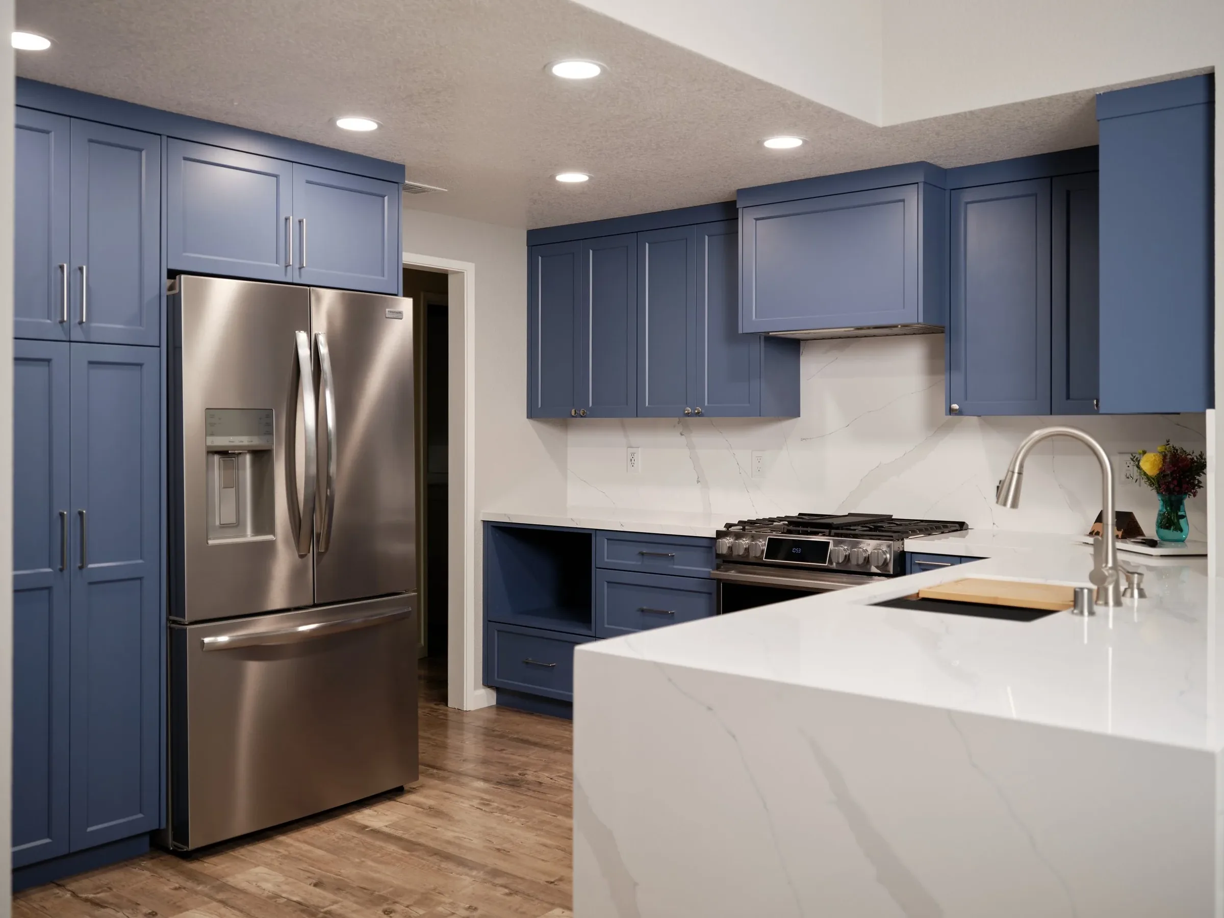 Modern kitchen with blue cabinets, stainless steel refrigerator, marble countertops, and a white island with a sink and faucet.