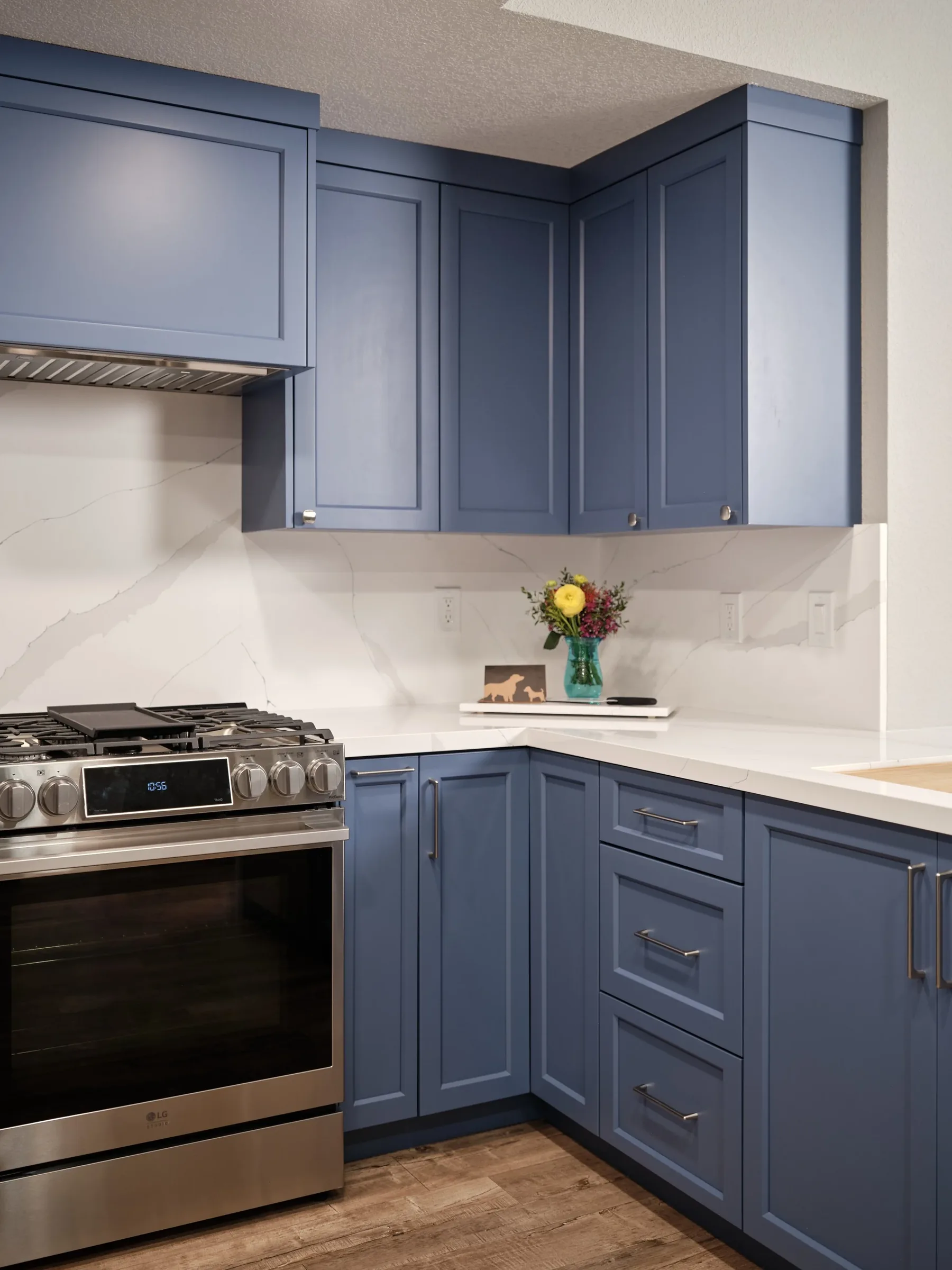 Kitchen with blue cabinets, stainless steel oven, white marble countertop, and a flower vase with colorful flowers.