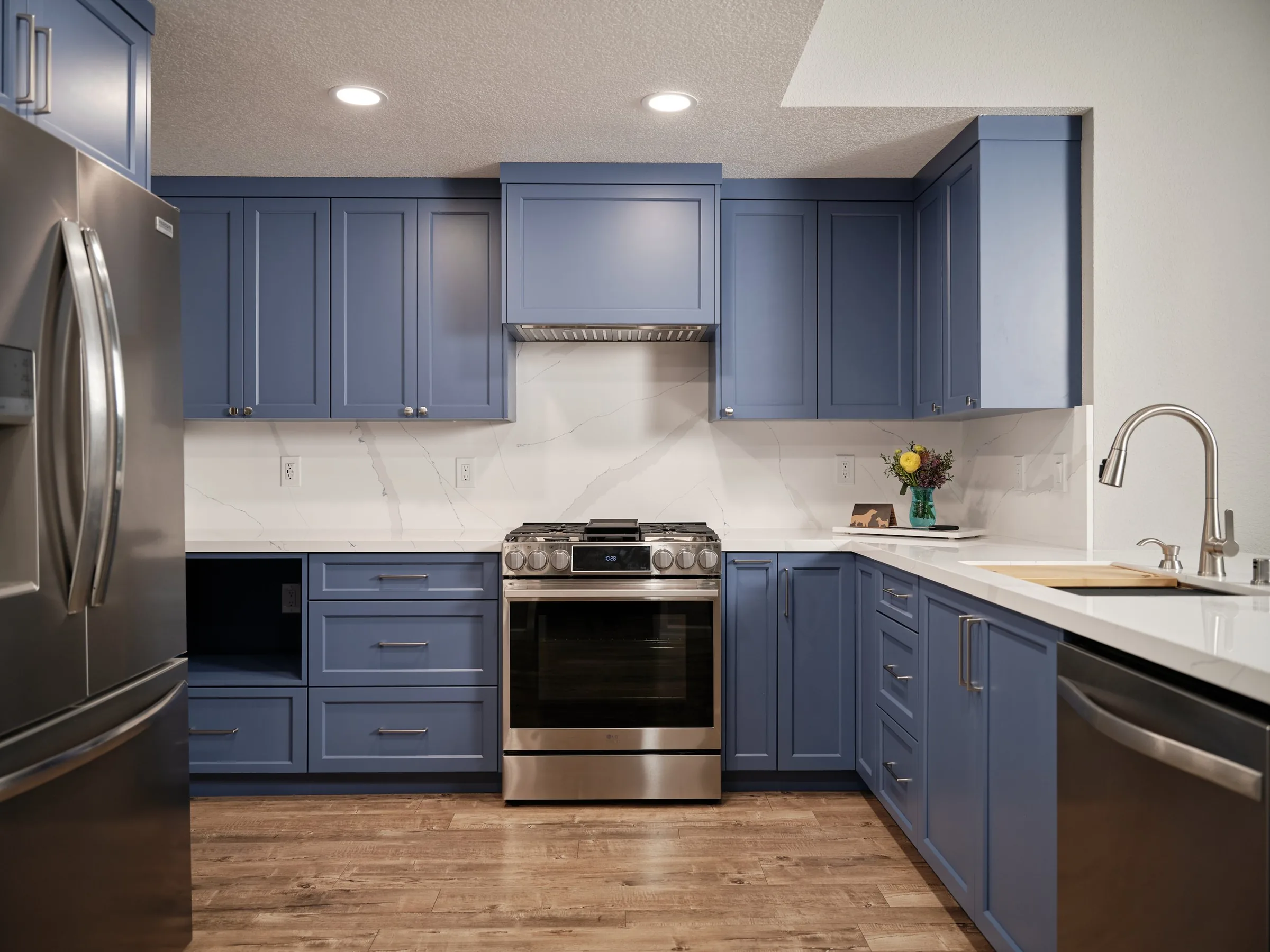 A modern kitchen with blue cabinets, a stainless steel stove, a marble backsplash, and a small floral arrangement on the counter.