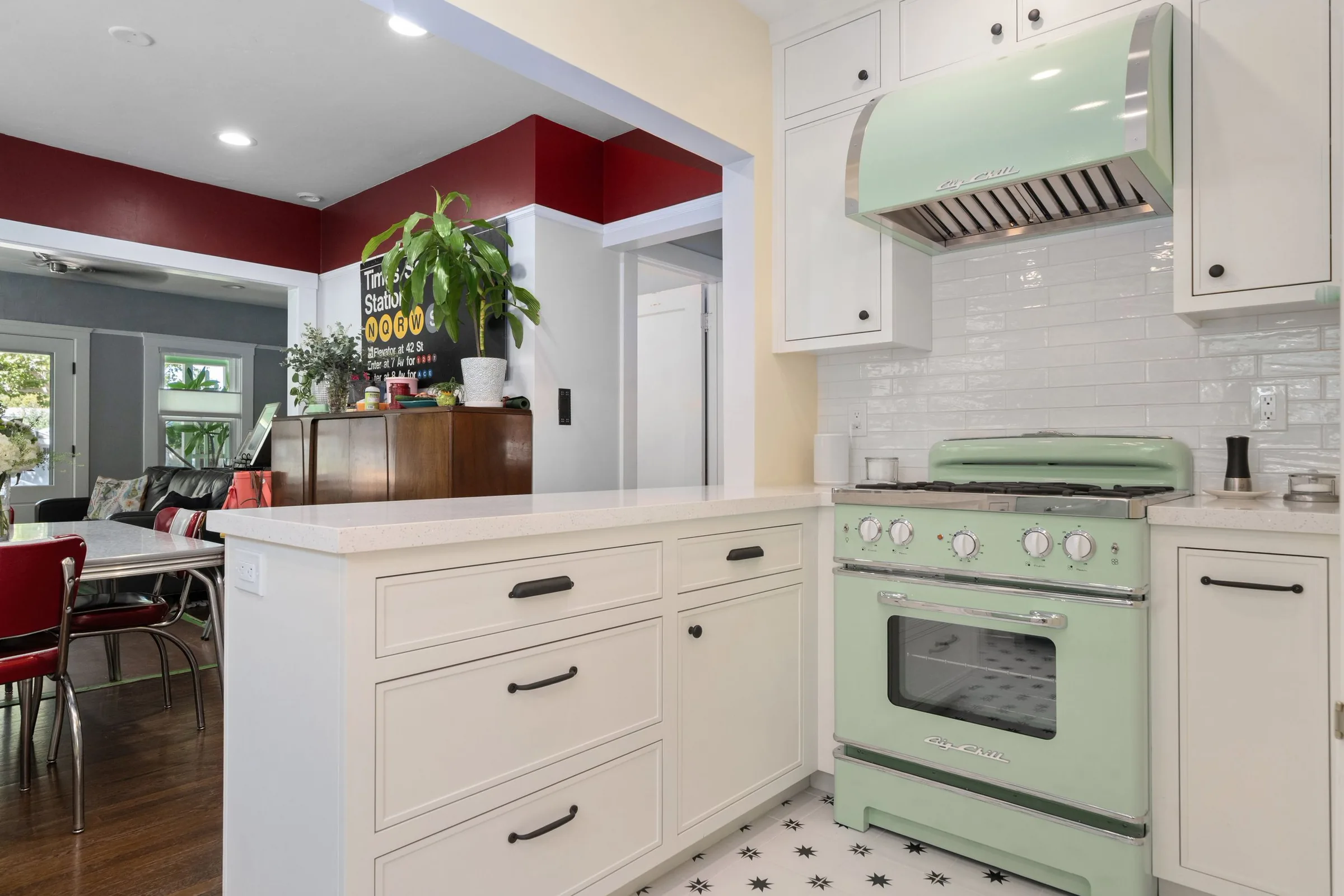 A kitchen with white cabinets and black knobs, a mint green vintage-style stove, and a white countertop. There are white subway tile backsplashes with black star pattern floor tiles in front of the stove. Some plants and a bulletin board are visible 