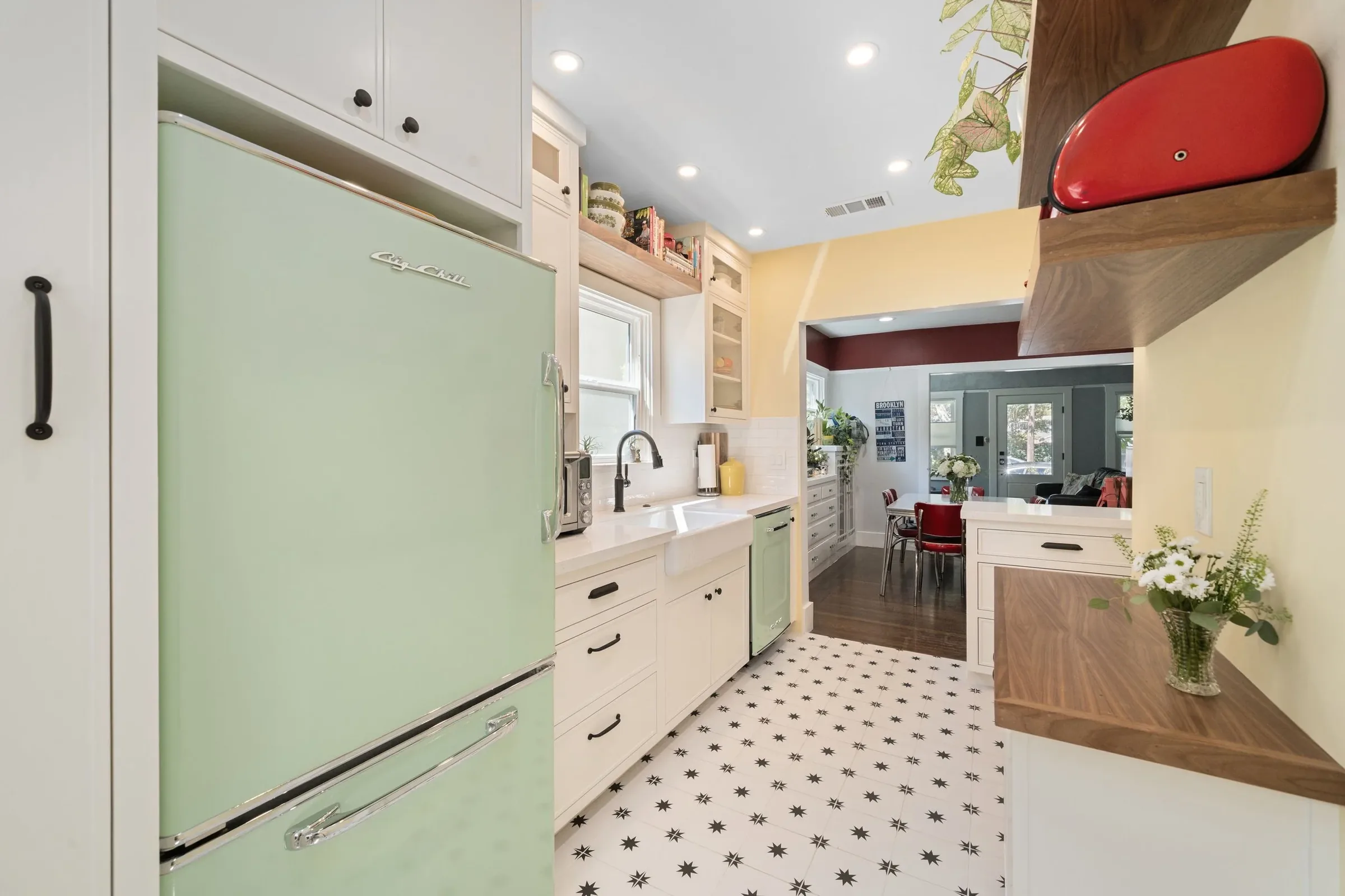 Kitchen with mint green retro refrigerator, white cabinets, black and white star patterned tile floor, open shelving with books and dishes, and a view into the dining and living areas with red chairs and a glass door.