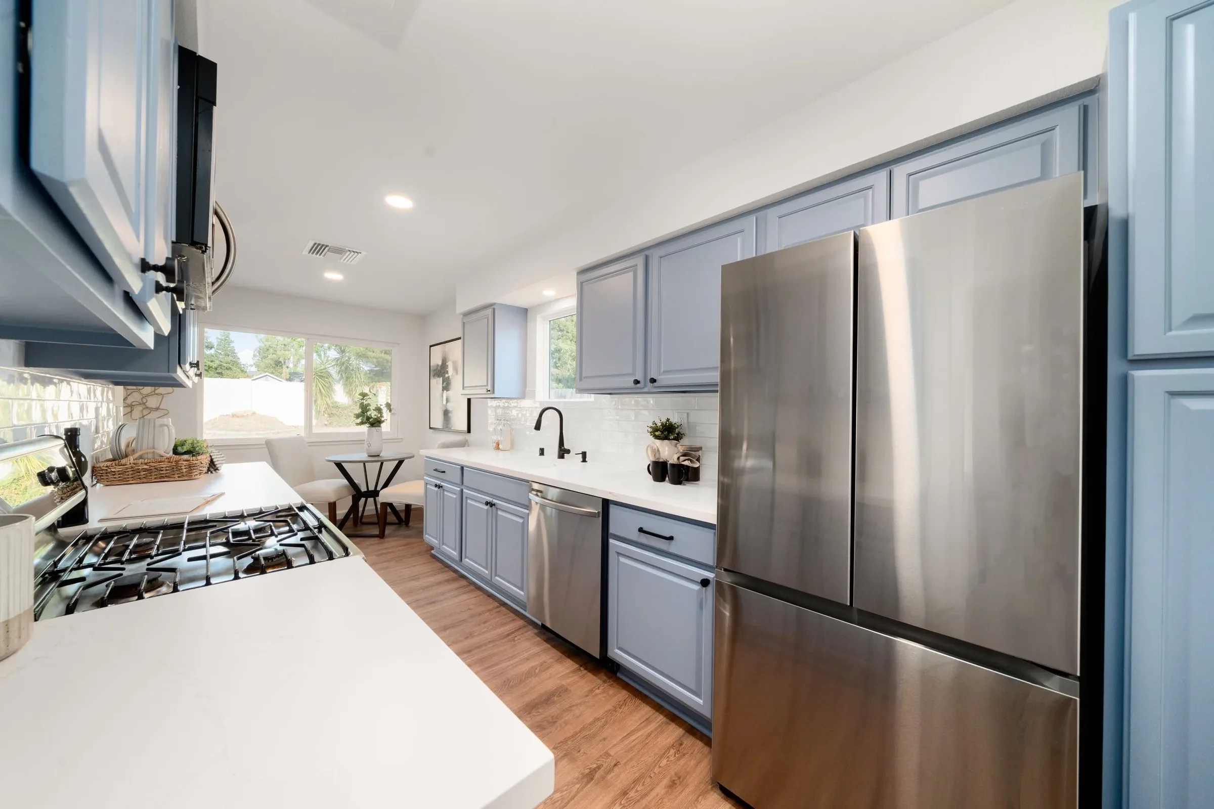 Modern kitchen with light blue cabinets, stainless steel appliances, white countertops, wood flooring, and a large window with outdoor view.