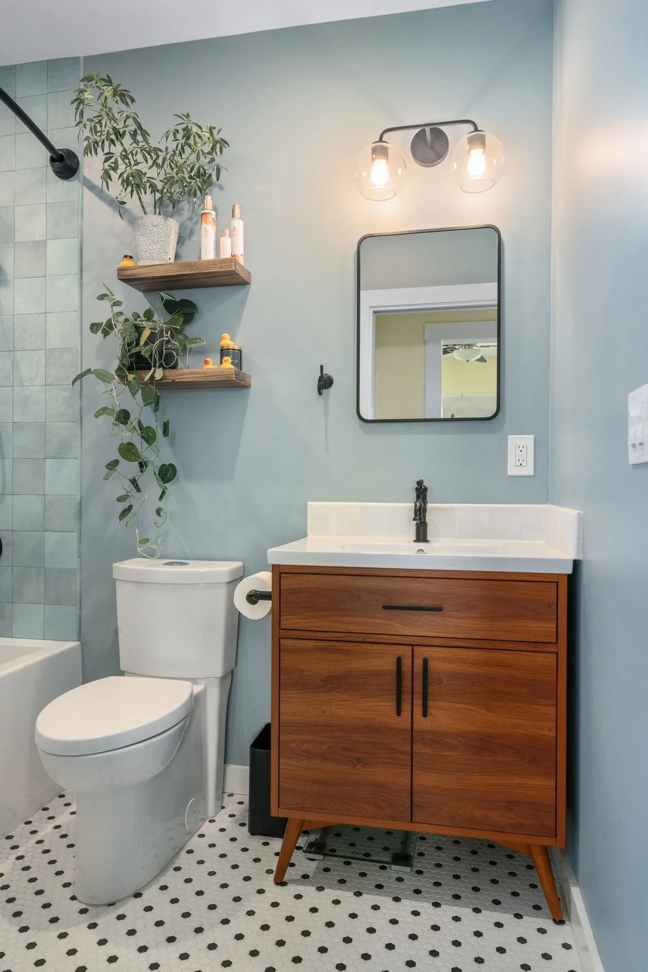 A modern bathroom with a white toilet, wooden vanity with black handles, wall mirror, wall-mounted light fixture, and two floating wooden shelves with potted plants and toiletries. The room has light blue walls and black and white checkered tile floo