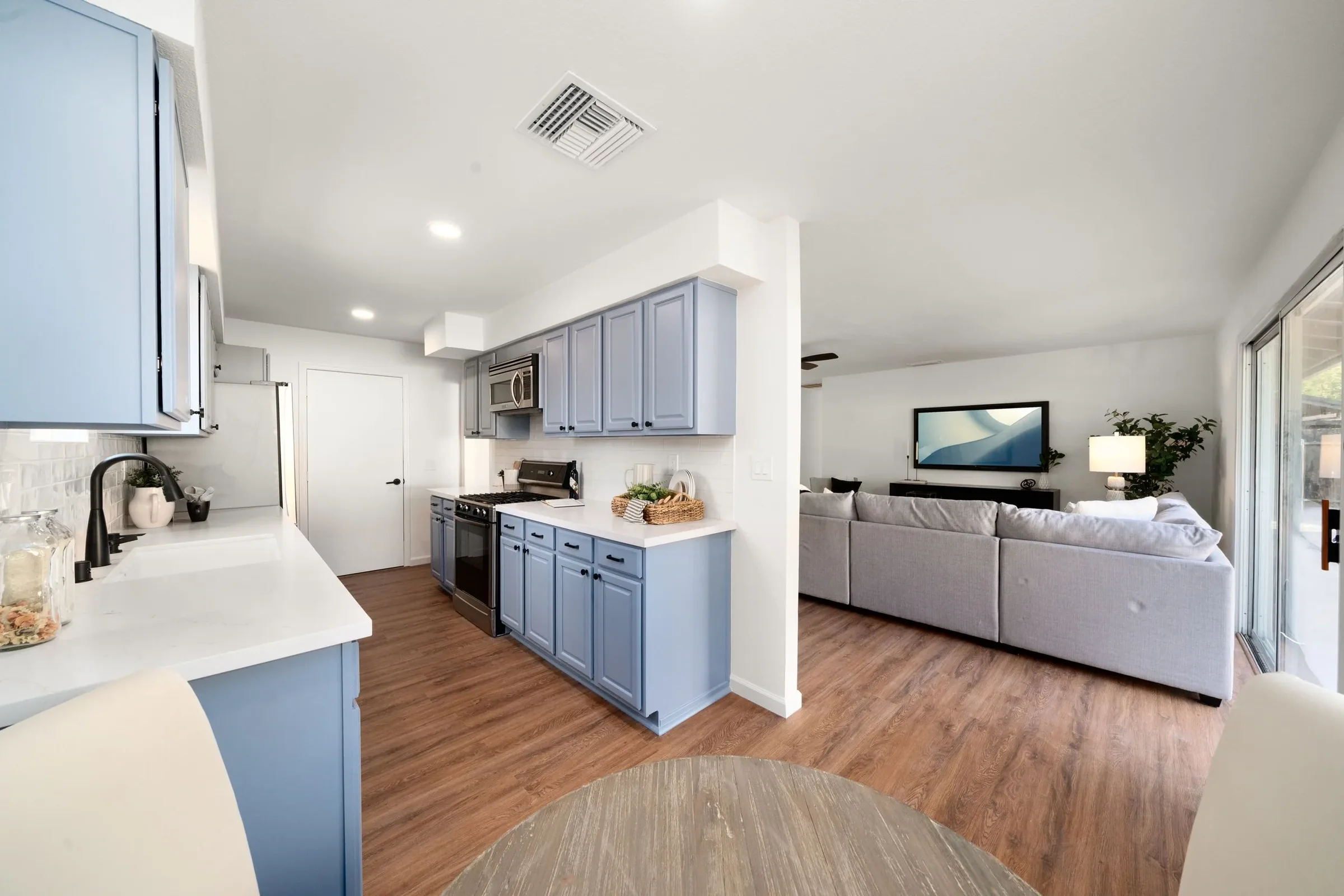 Open-concept kitchen and living room with blue cabinets, white walls, a gray sofa, wall-mounted TV, and large windows with sliding glass doors.