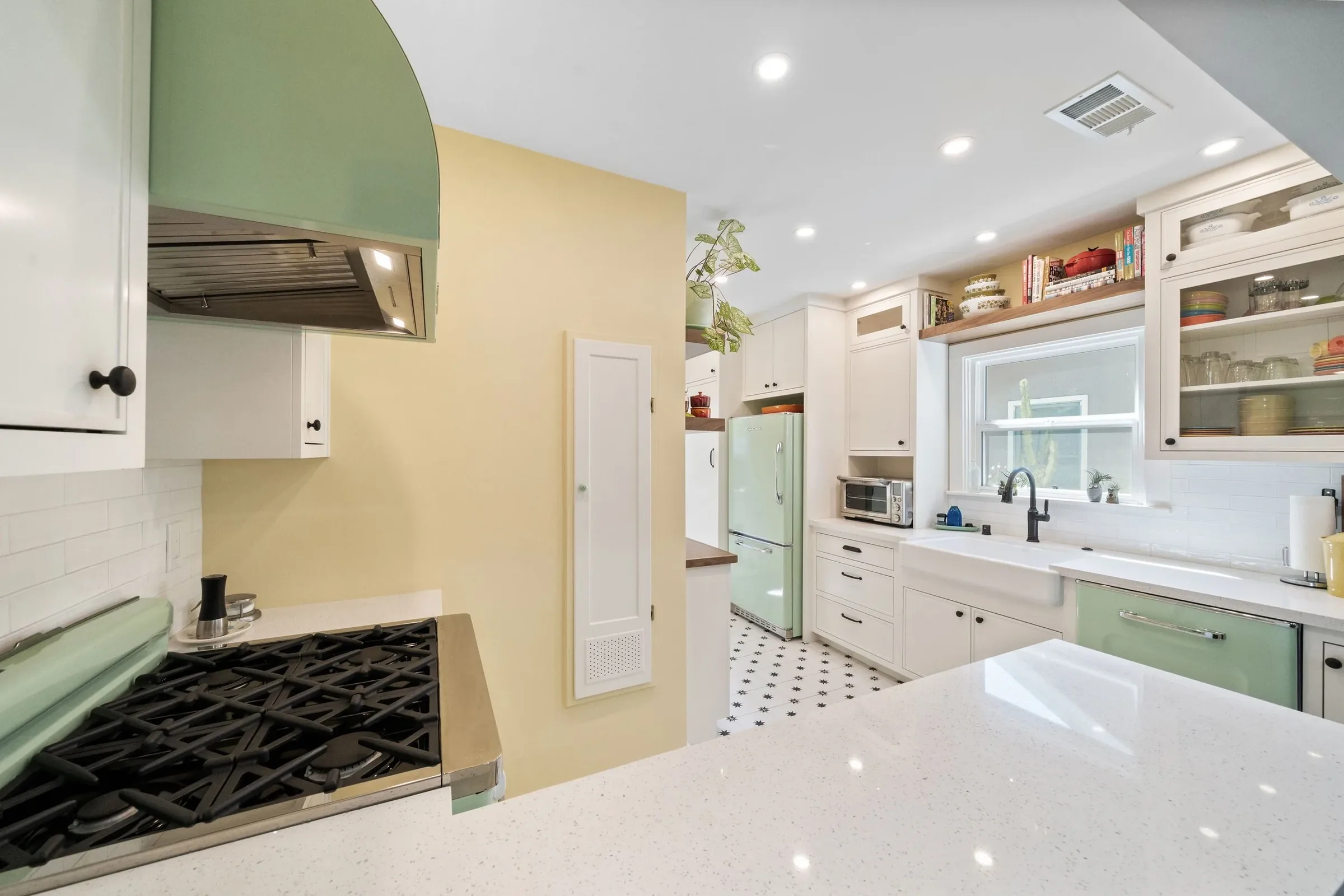 A bright kitchen with white cabinets, black hardware, a light countertop, a stove with a green range hood, a window above the sink, open shelves with dishes and cookbooks, a small refrigerator, and decorative items, with a yellow wall and black-and-white patterned floor.