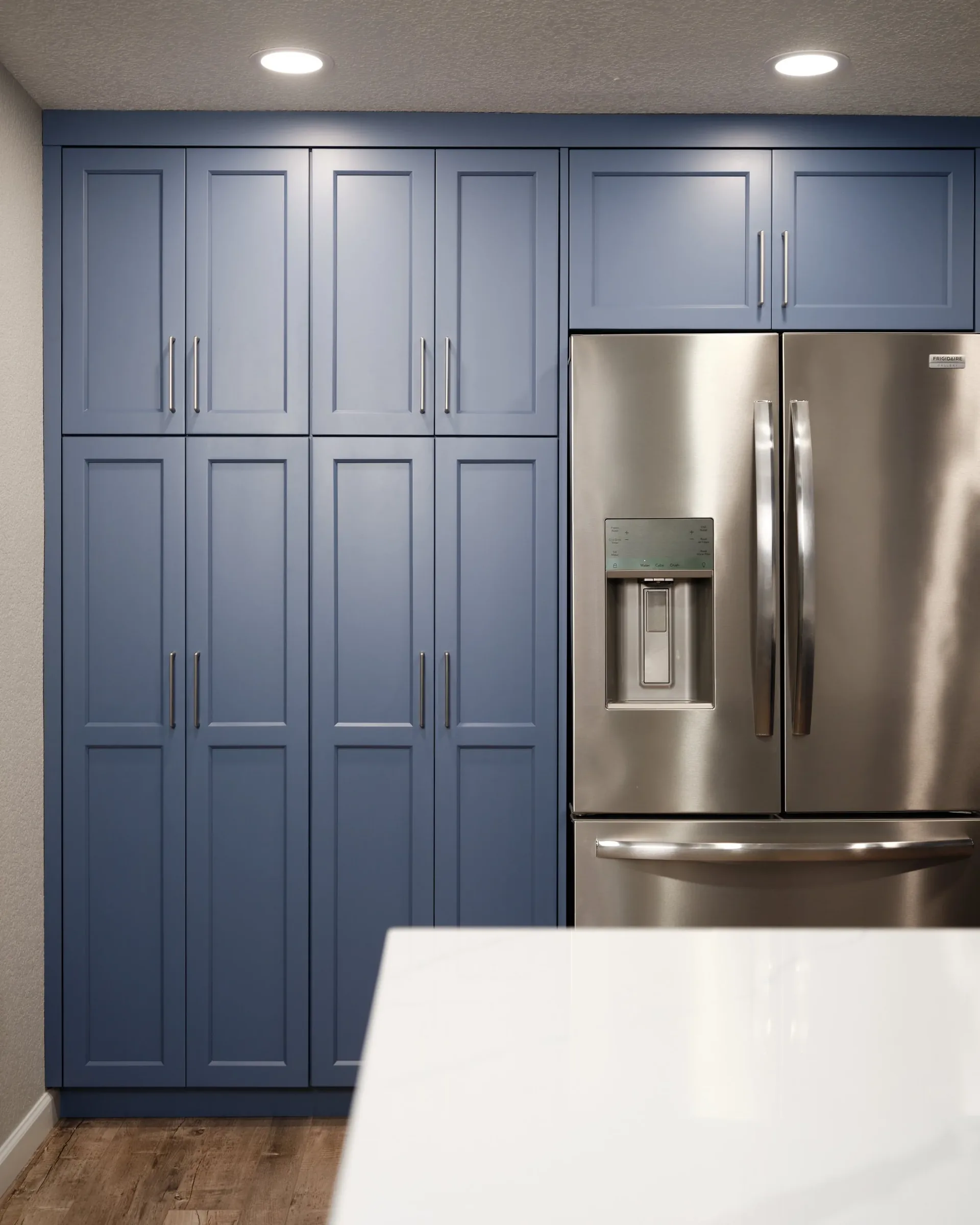 Blue kitchen cabinets with stainless steel Frigidaire Gallery refrigerator. White countertop in foreground, brown wood floor, gray walls, ceiling with recessed lighting.