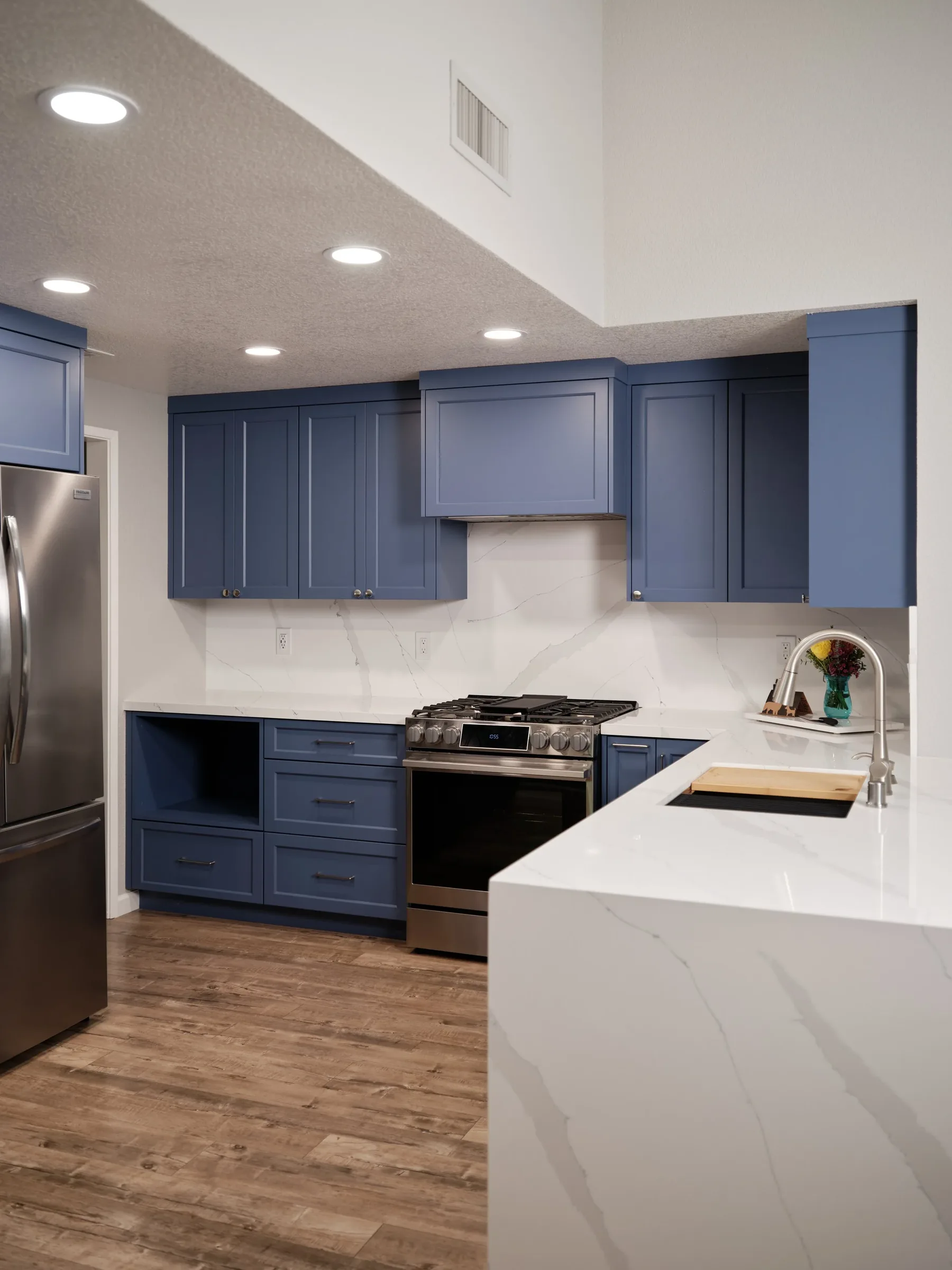 Modern kitchen with blue cabinets, stainless steel refrigerator and oven, white marble countertops, and dark hardwood floors.