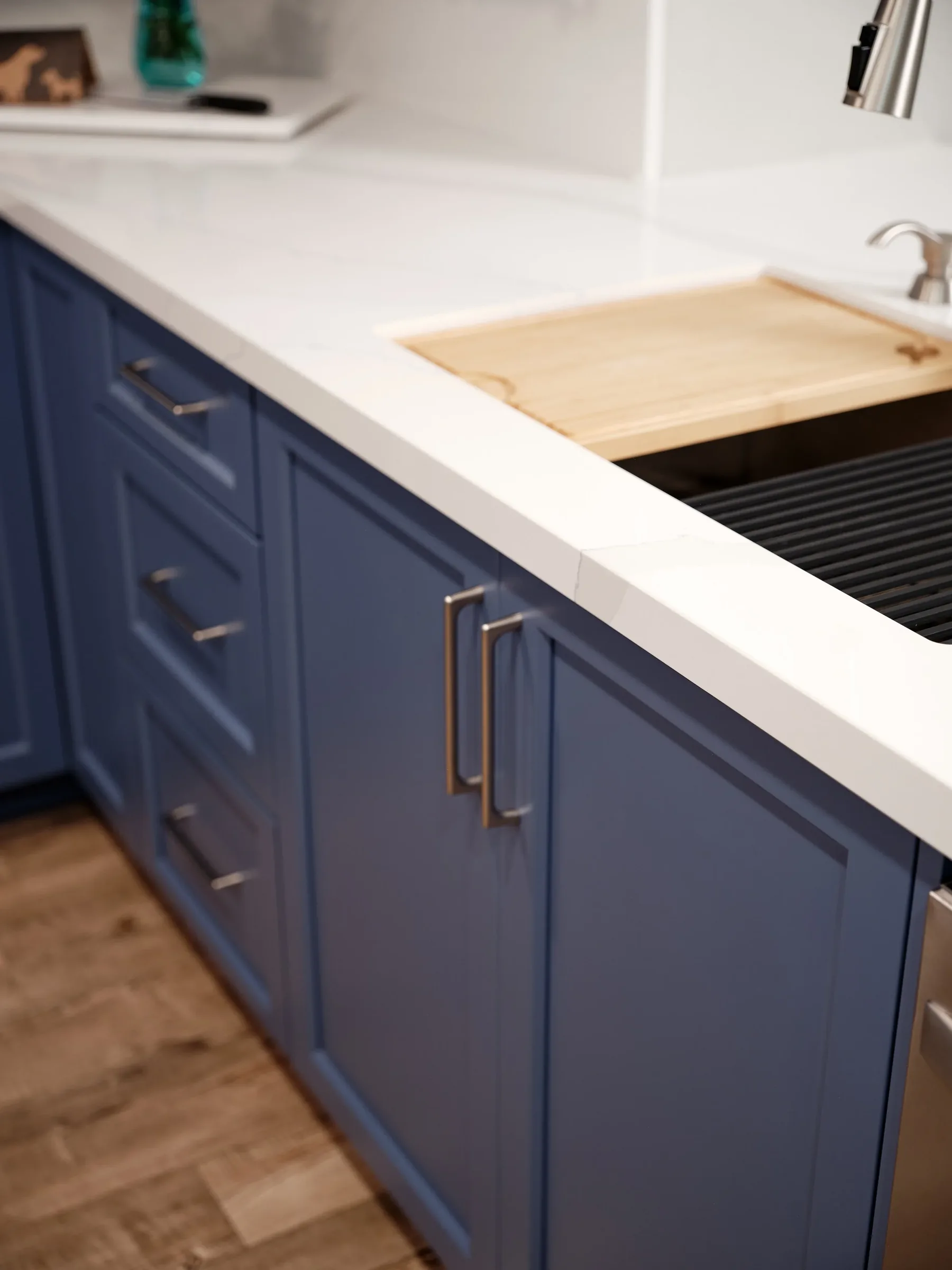 A kitchen cabinet with a blue finish, silver handles, and a white countertop. There is a wooden cutting board and a black dish drainer. The floor is wooden.